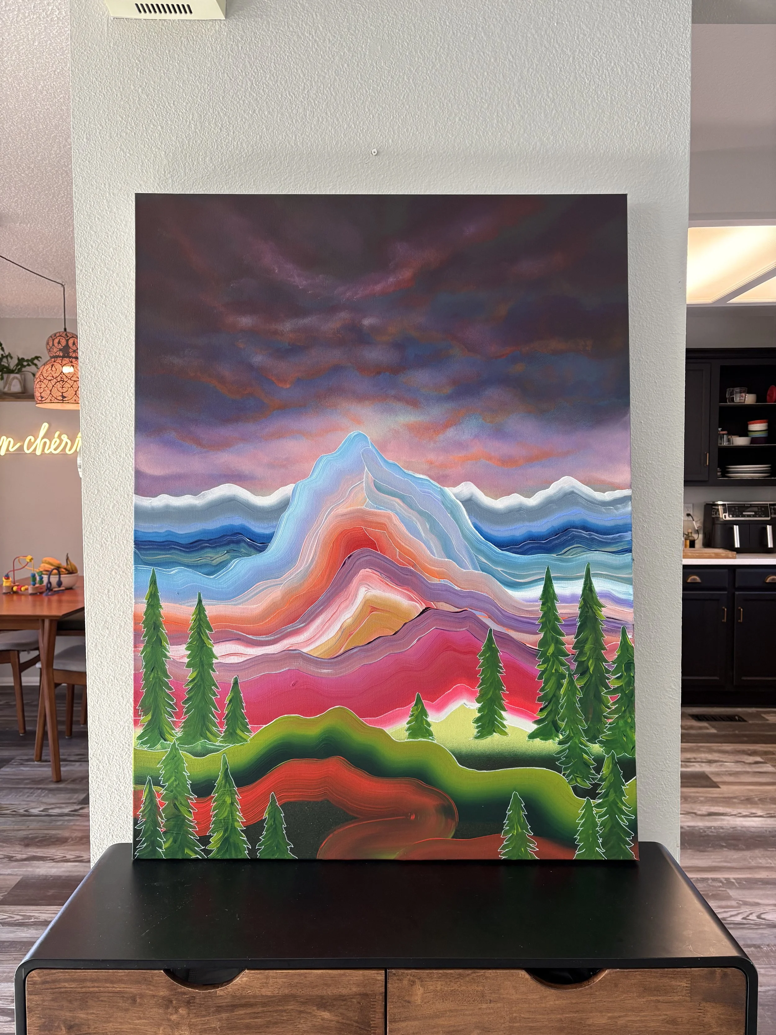 Even Under Dark Skies painting by Colorado artist Jessie Belisle — vibrant layered mountain landscape beneath a dark dramatic sky with evergreen trees.