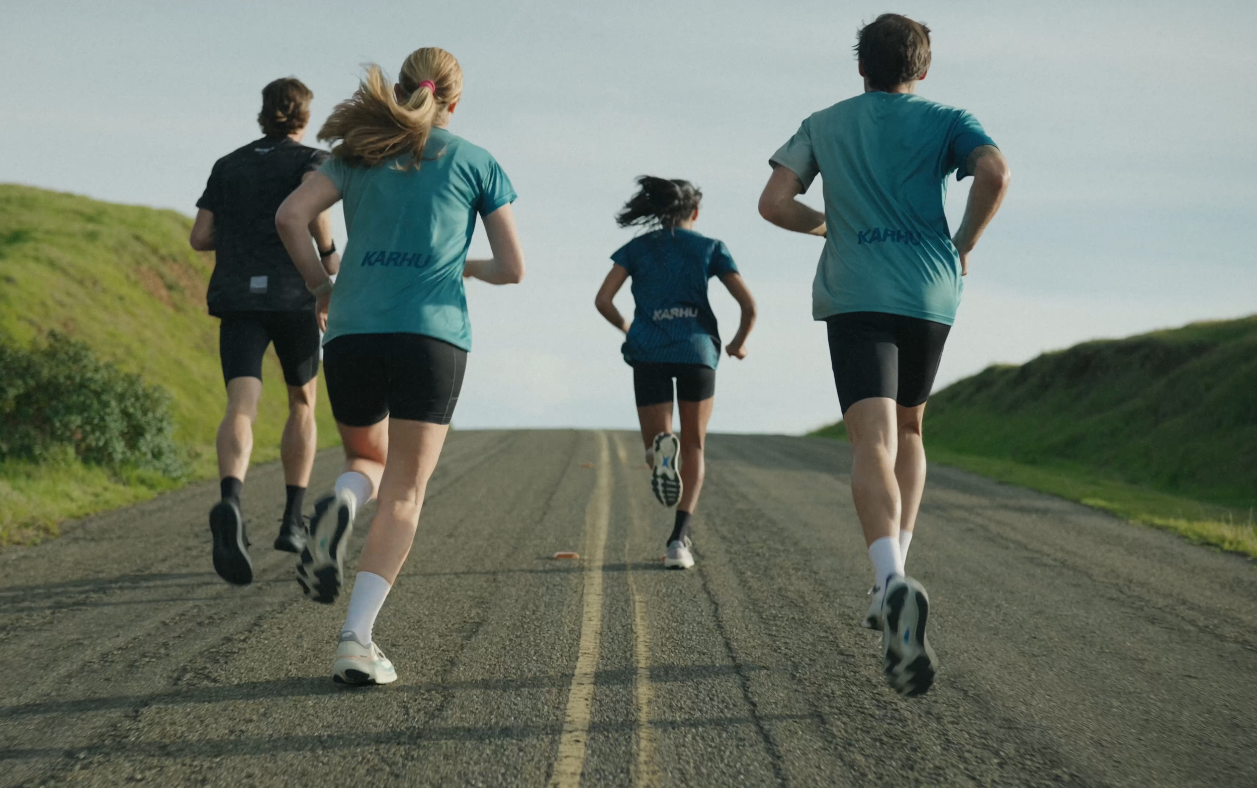 Four people running on a road in a rural area, with green hills on the sides, during daylight.