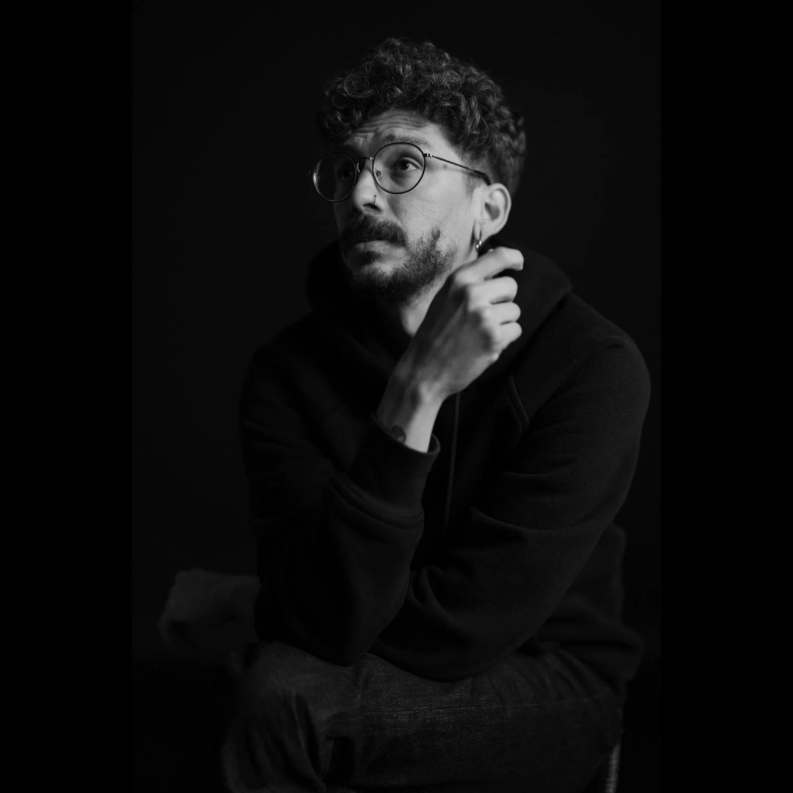 Black and white portrait of a young man with curly hair and glasses, wearing a hoodie, looking upward with his hand on his neck, against a dark background.