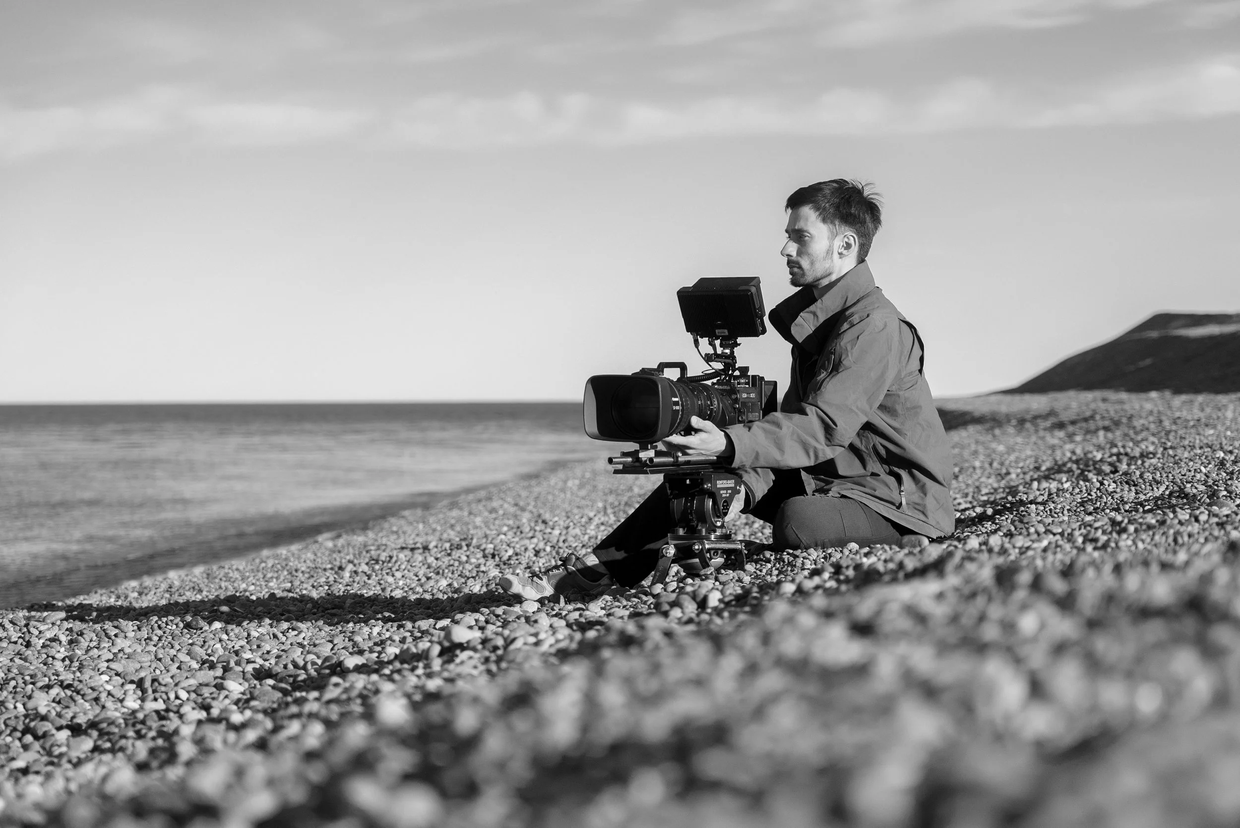 A man sitting on a pebble beach, operating a professional video camera mounted on a tripod, with the ocean and a hill in the background, in black and white.