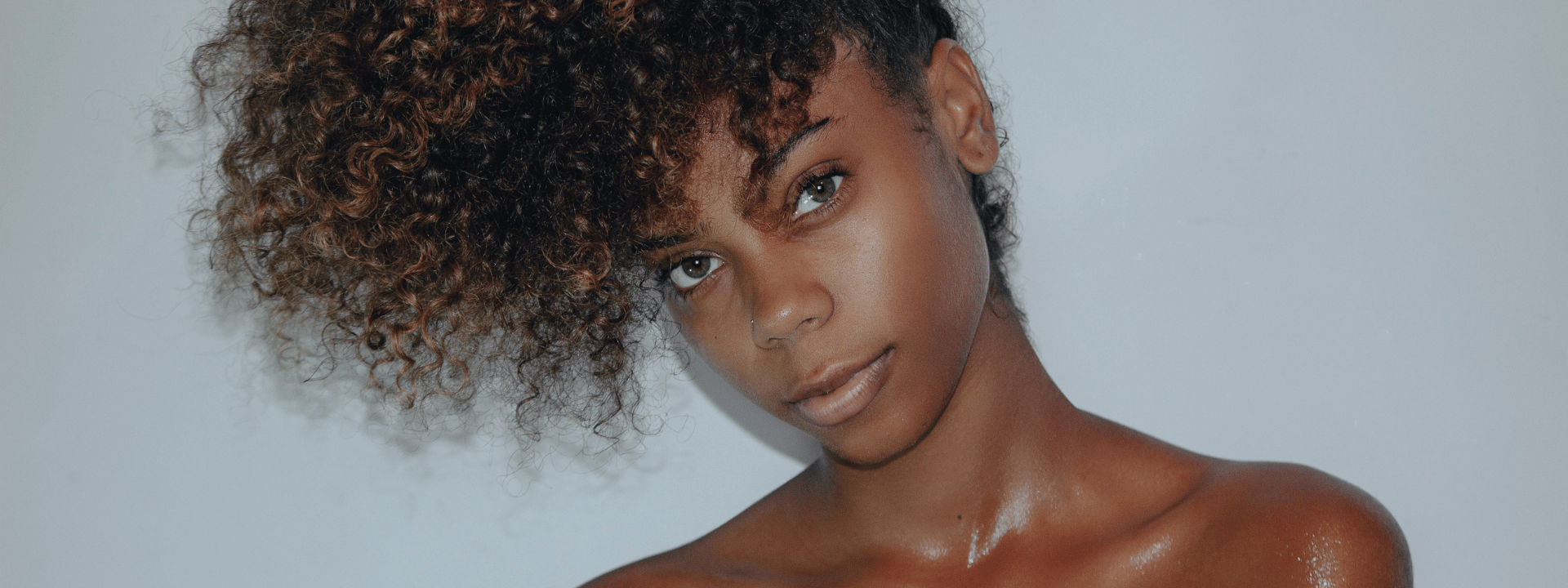 Close-up portrait of a young woman with natural curly hair, light brown skin, and subtle makeup, looking at the camera against a plain, light-colored background.