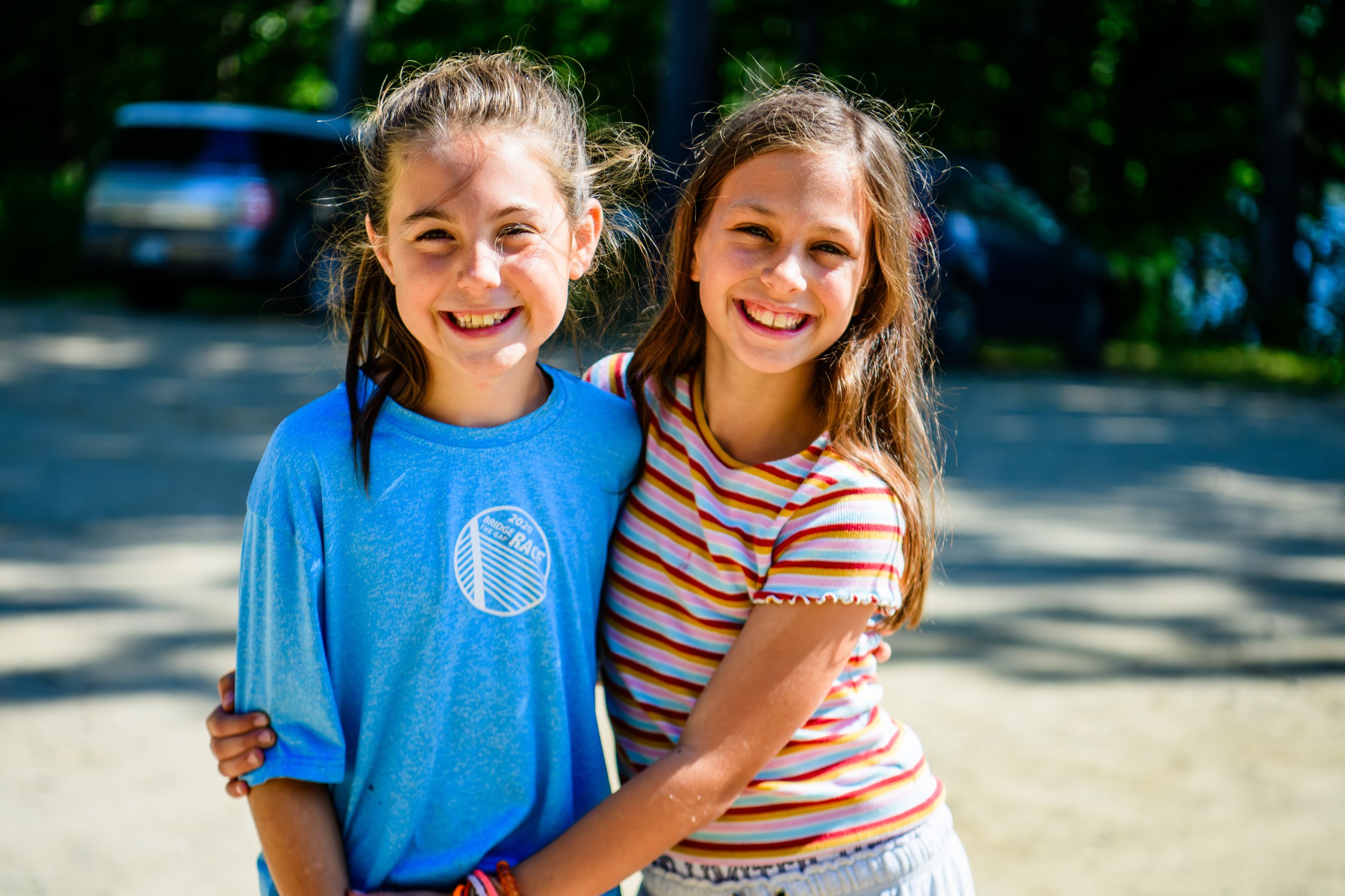 Two young girls smiling and hugging outdoors on a sunny day, with cars and trees in the background at Kingdom Life Church Kingdom Kids in Oakland, Maine.