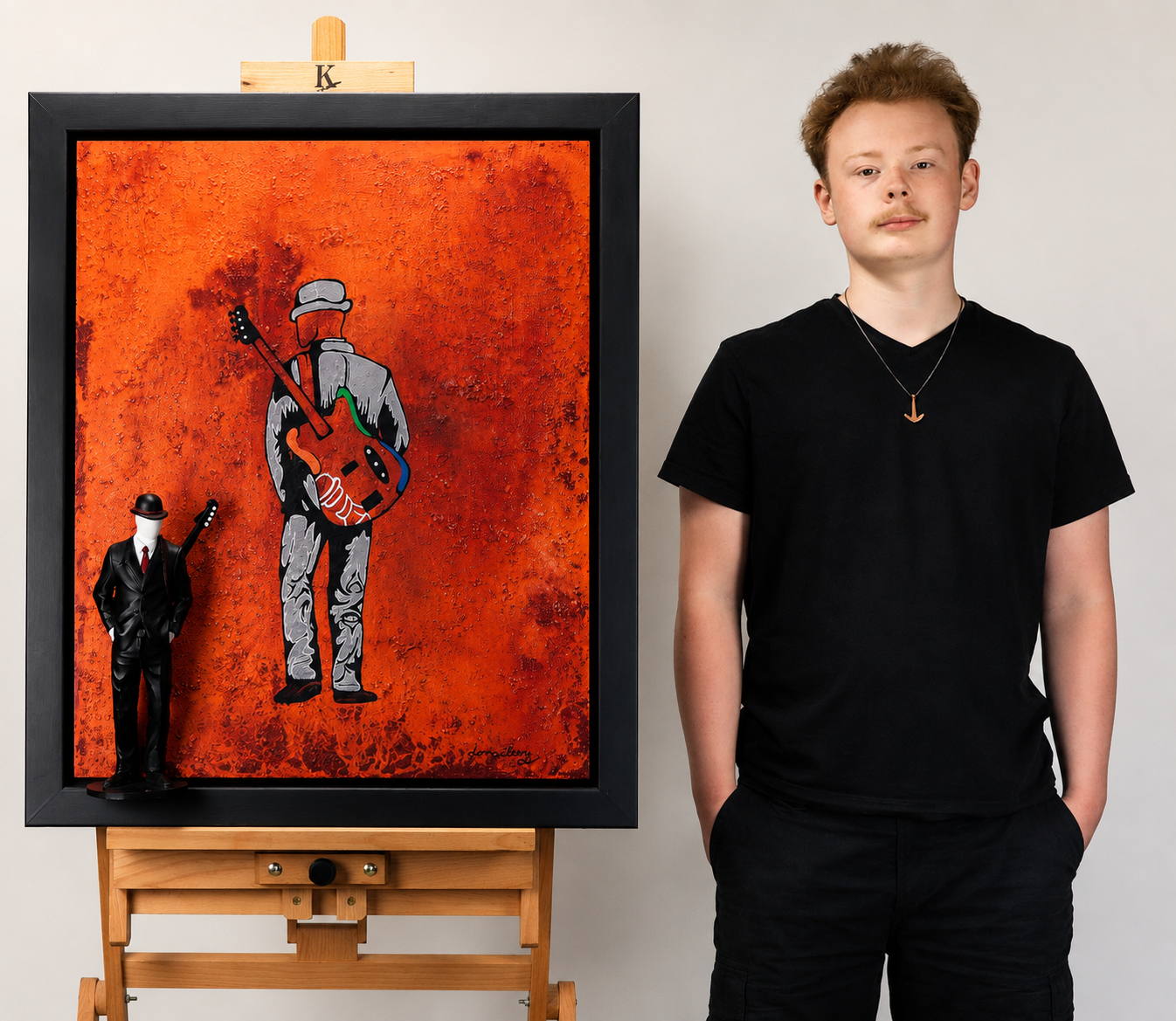 En ung mann står ved siden av et kunstverk med en graffiti-stil av en person med gitar på en oransje bakgrunn. Kunstverket inkluderer også en skulptur av en mann i dress og hatt som holder en gitar.