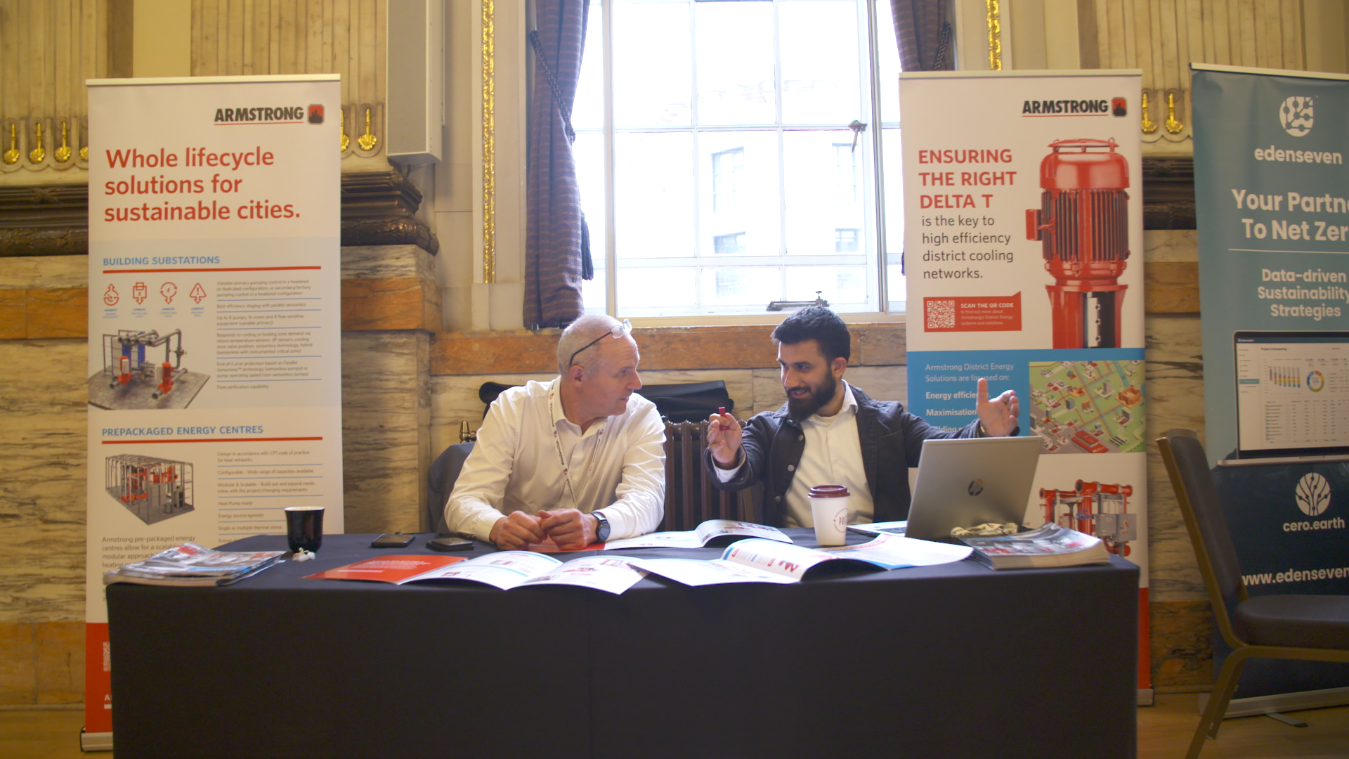 Two men sitting at a table talking at a conference booth with informational banners about energy solutions.