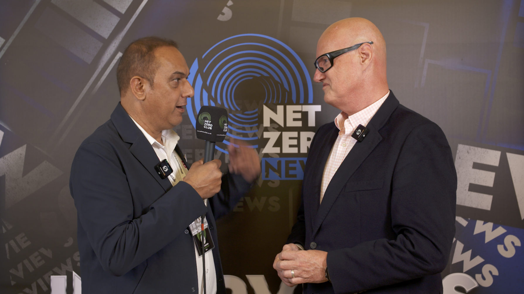 Two men in suits having an interview at a news event, with a backdrop displaying the words 'NET ZERO NEWS' and graphics related to news and media.
