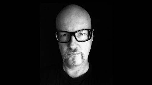A black-and-white portrait of a bald man wearing glasses, with a serious expression on his face.