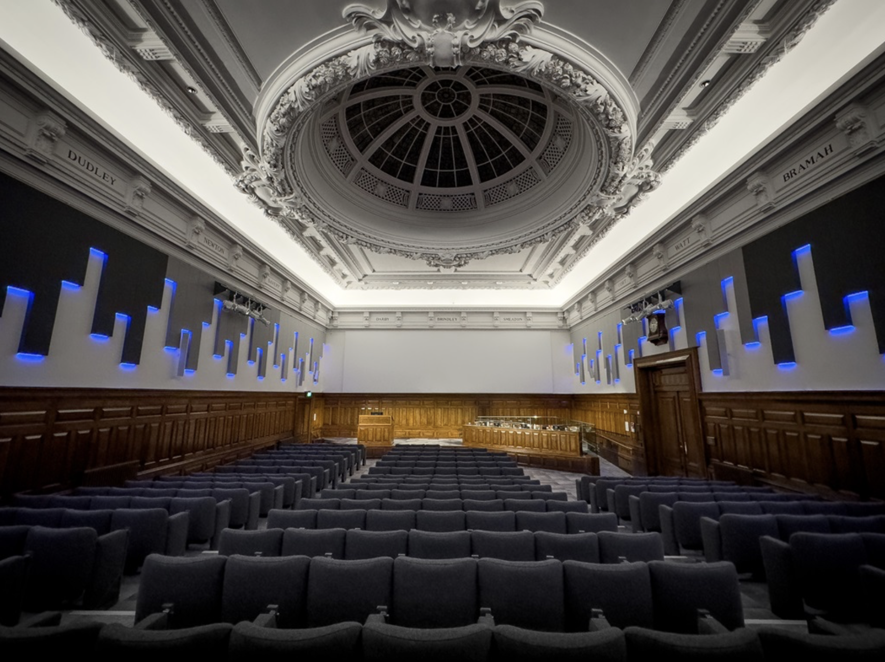 Empty auditorium with rows of black chairs, a wooden stage, and wall-mounted blue lights. Ornate ceiling with intricate moldings and a large round skylight.