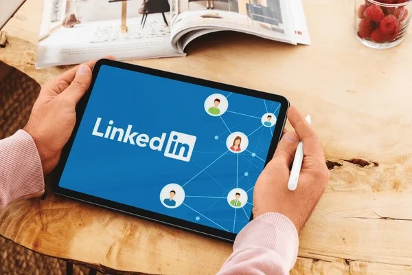 Person holding a tablet displaying the LinkedIn logo and network connection icons, with a notebook and a glass jar of raspberries on a wooden table.