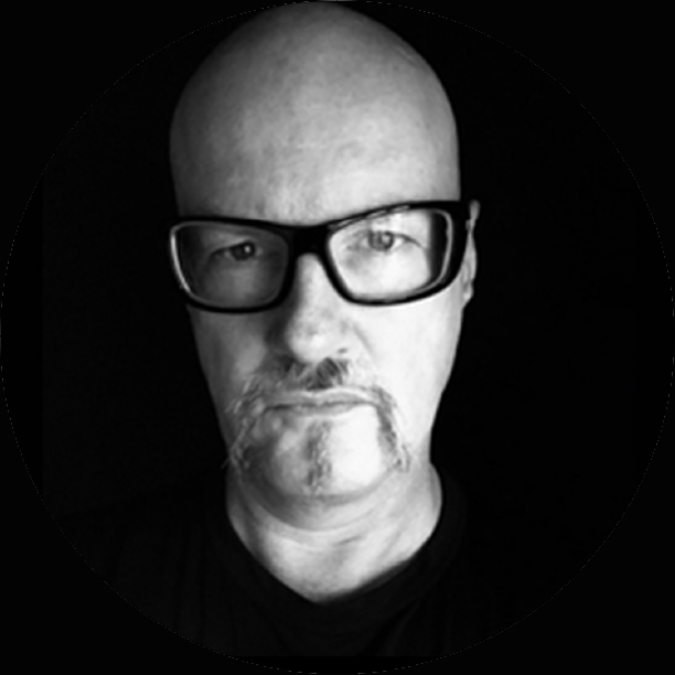 Black and white close-up photo of a bald man wearing glasses and a mustache with a goatee, looking directly at the camera.