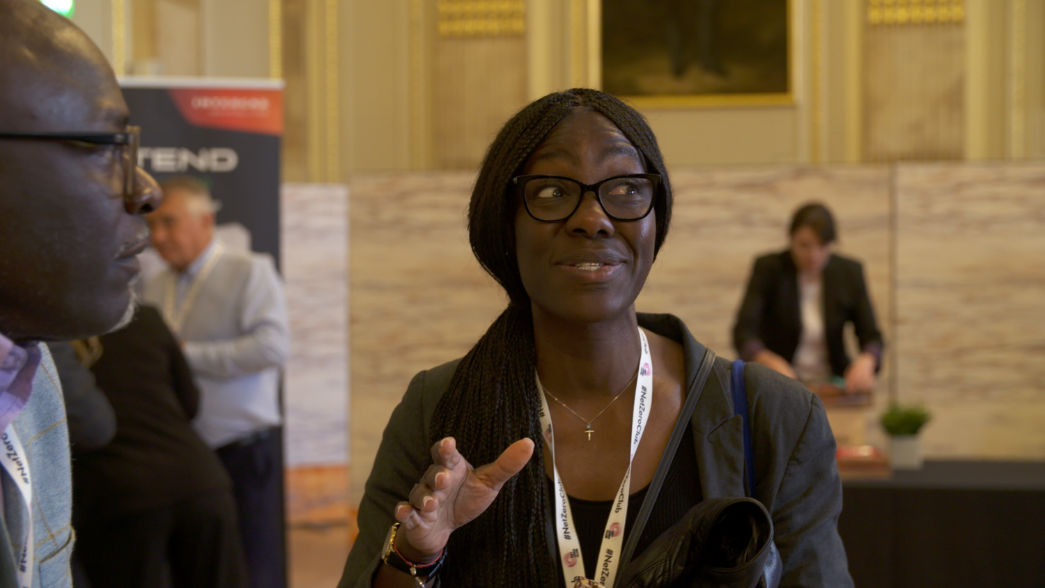 A woman with glasses and long braided hair speaking and gesturing with her hand in a conference or networking event with other attendees in the background. — London 2025.
