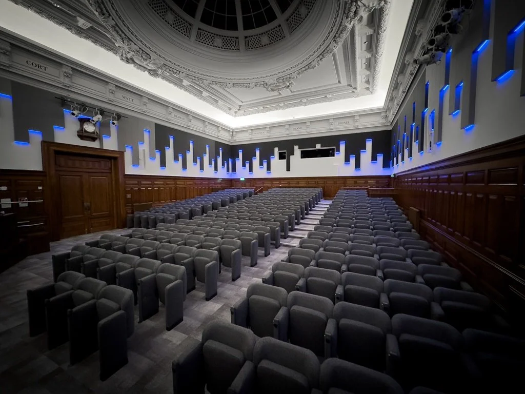 Empty auditorium with rows of gray chairs, wooden paneling, ceiling with ornate design, and blue accent lighting on the walls.