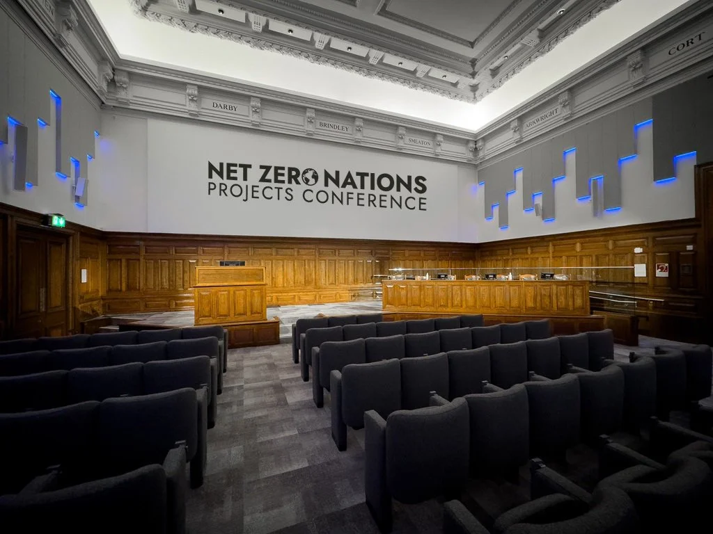 Empty conference room with wooden paneling, chairs arranged facing a stage with a podium, and a sign that reads 'NET ZERO NATIONS PROJECTS CONFERENCE' on a white wall.