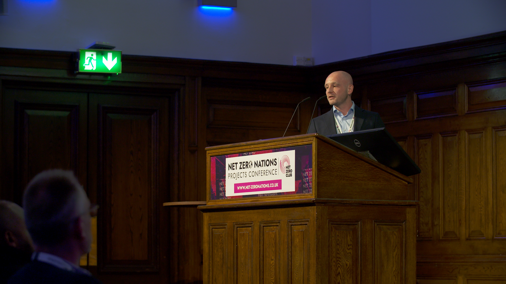 A man giving a presentation at the NET ZERO NATIONS Projects Conference, standing behind a wooden podium with a sign showing the event's branding and website. The conference is indoors with dark wood paneling and an emergency exit sign overhead.