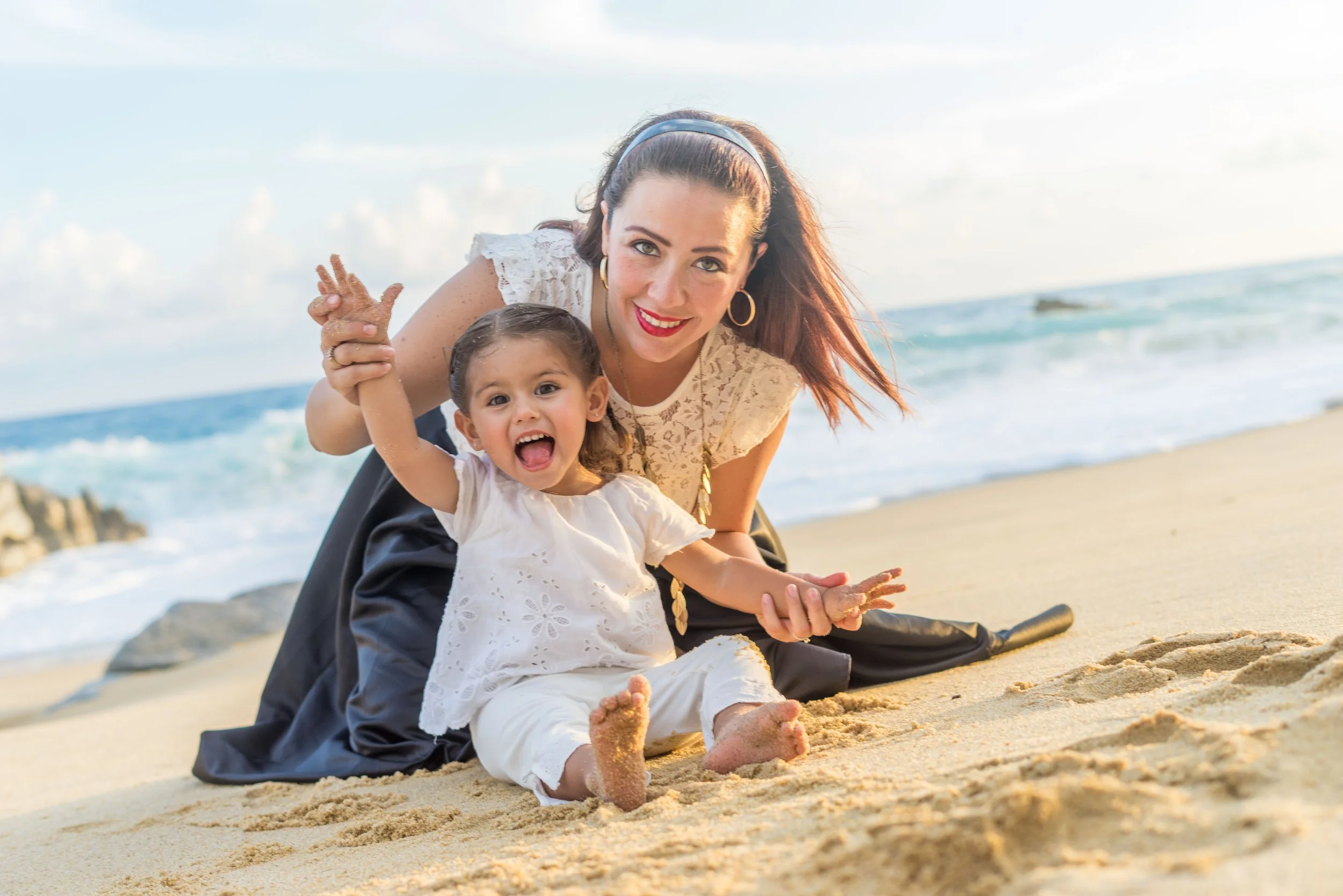 A woman and a young girl playing and sitting on the sandy beach near the ocean, smiling and with sand on their hands and feet.