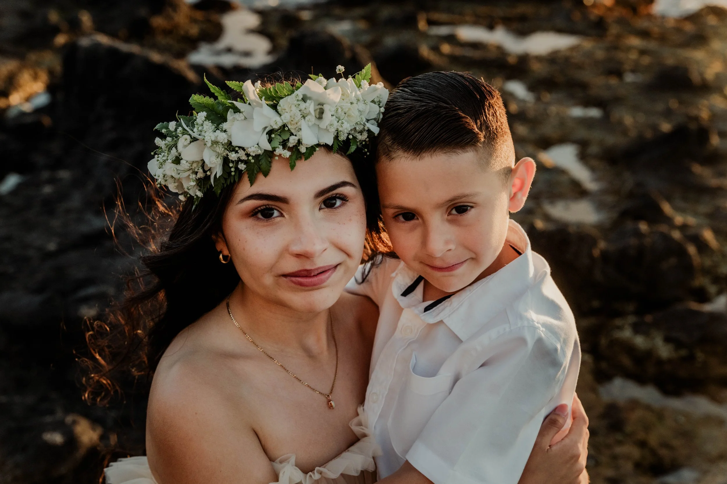 A woman wearing a white floral headband holding a young boy at sunset by the rocks on the beach.