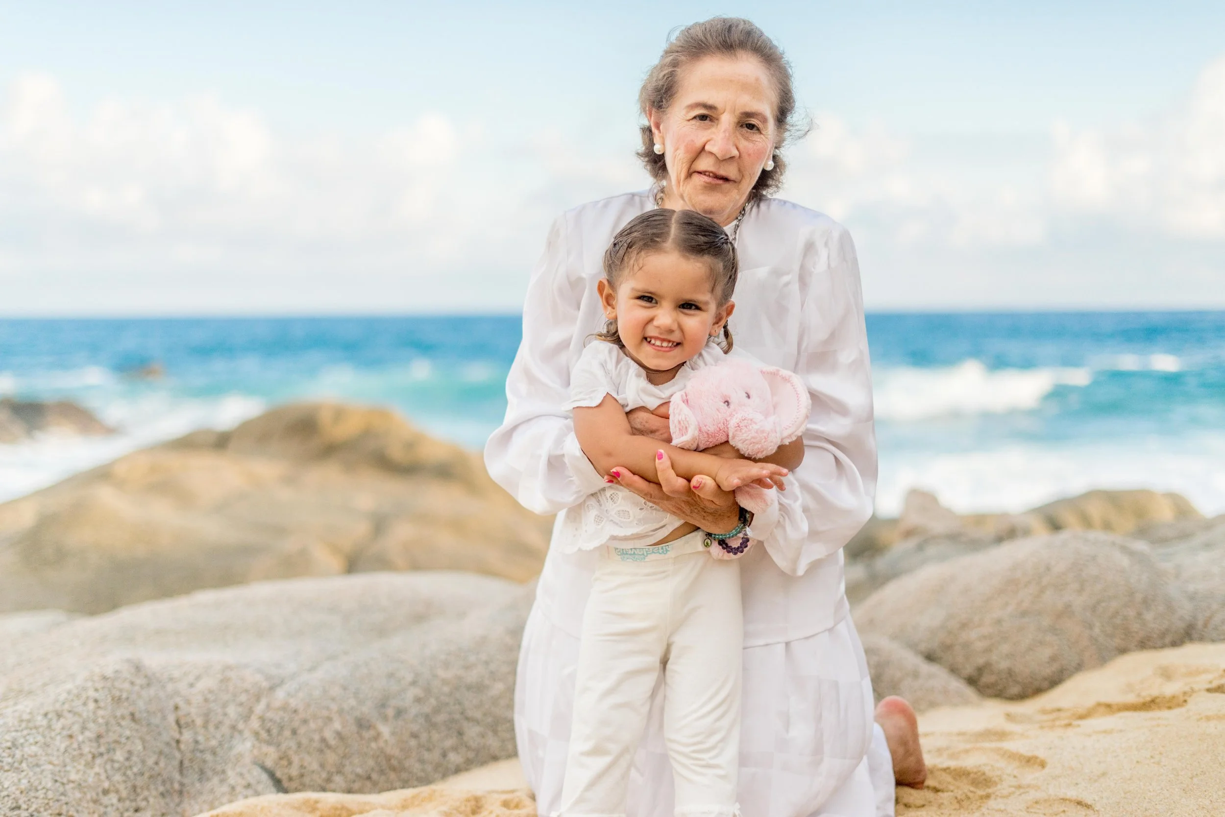 An elderly woman and a young girl kneeling on a beach with rocks and ocean waves in the background. The woman is holding the girl who is holding a pink stuffed animal.
