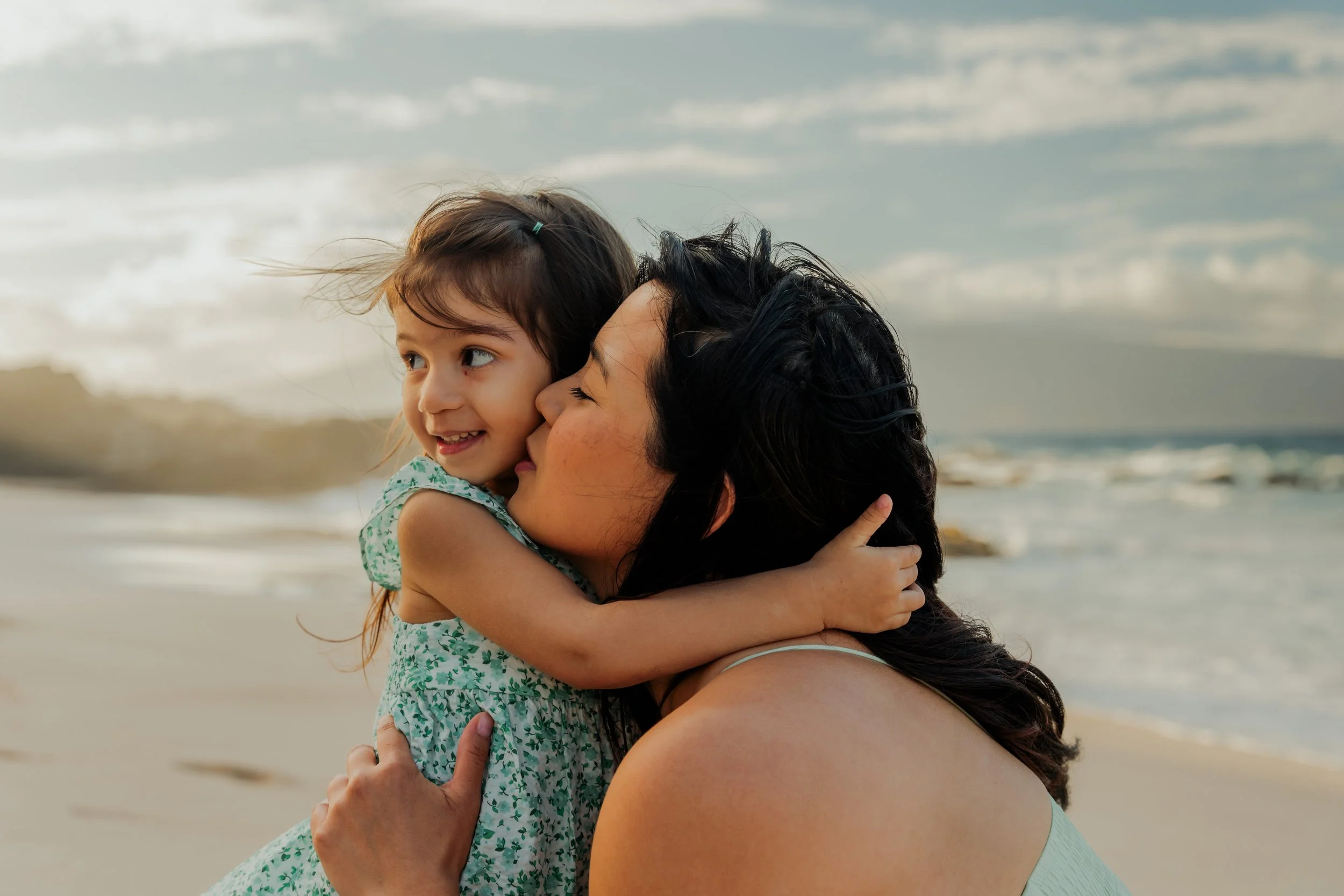 A woman and a young girl embrace and share a kiss at the beach, with ocean waves and cloudy sky in the background.