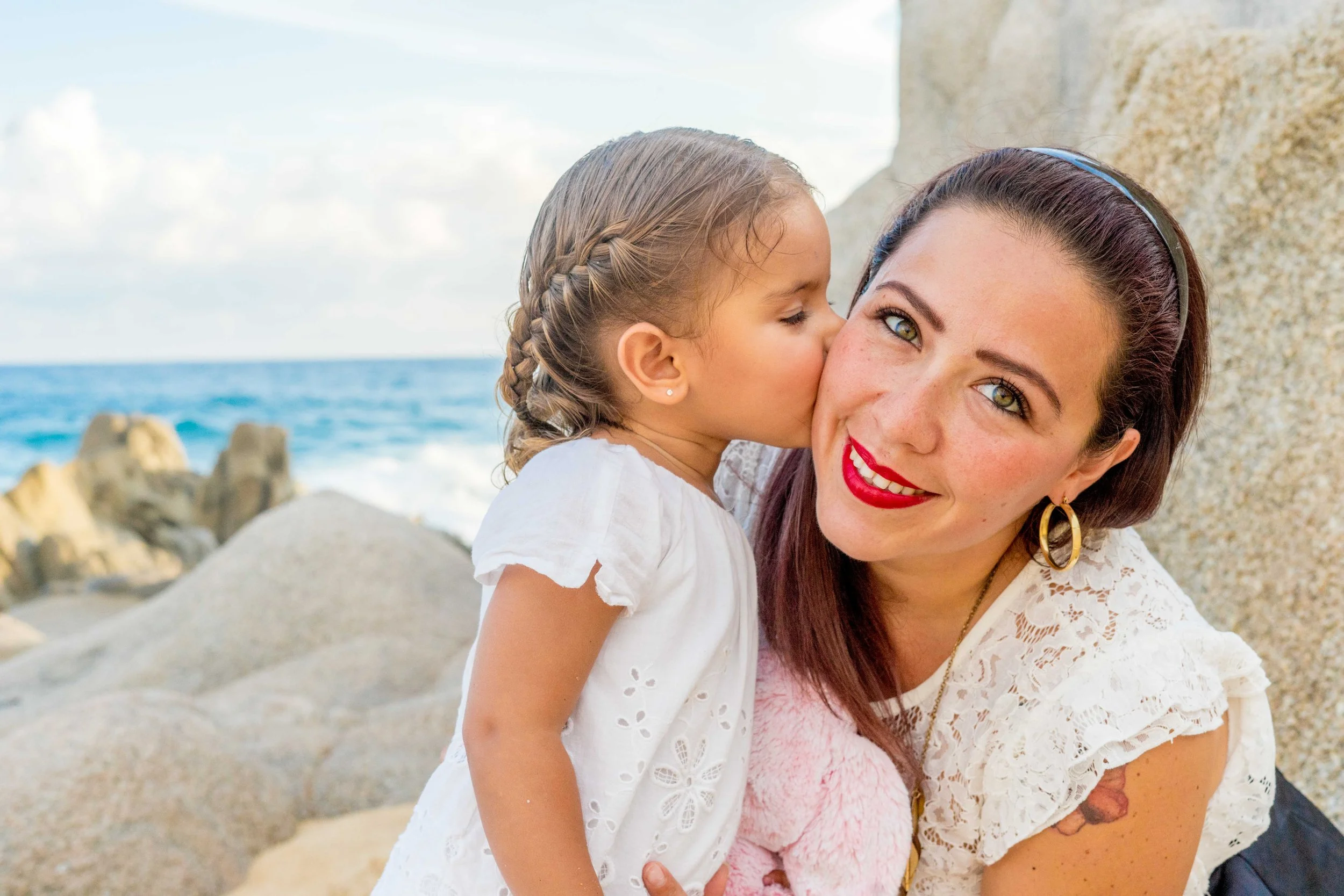 A woman with red lipstick, earrings, and a headband kisses a young girl on the cheek near the beach, with rocks and the ocean in the background.
