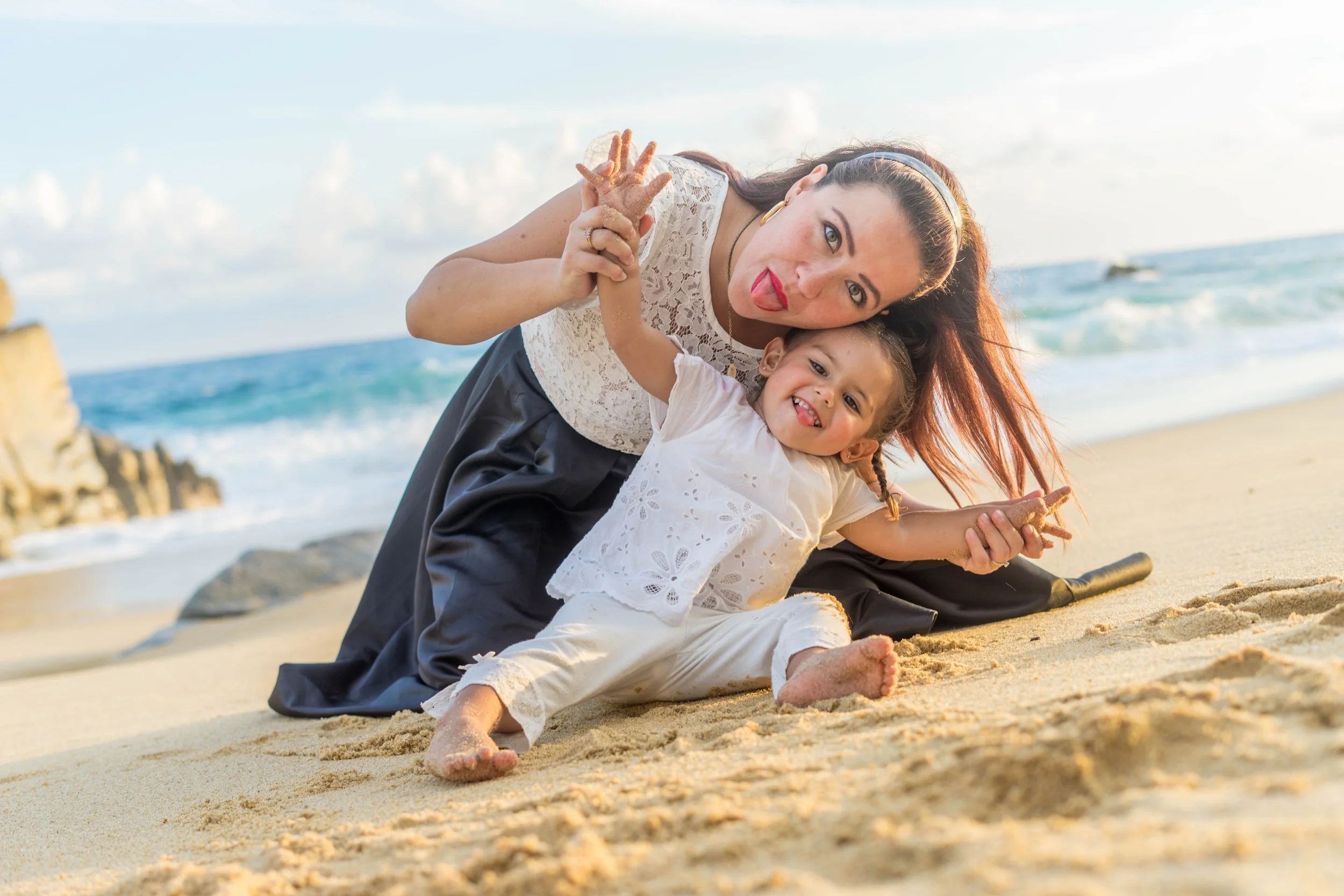 Woman and young girl playing on a sandy beach near the ocean, smiling and making funny faces at the camera.