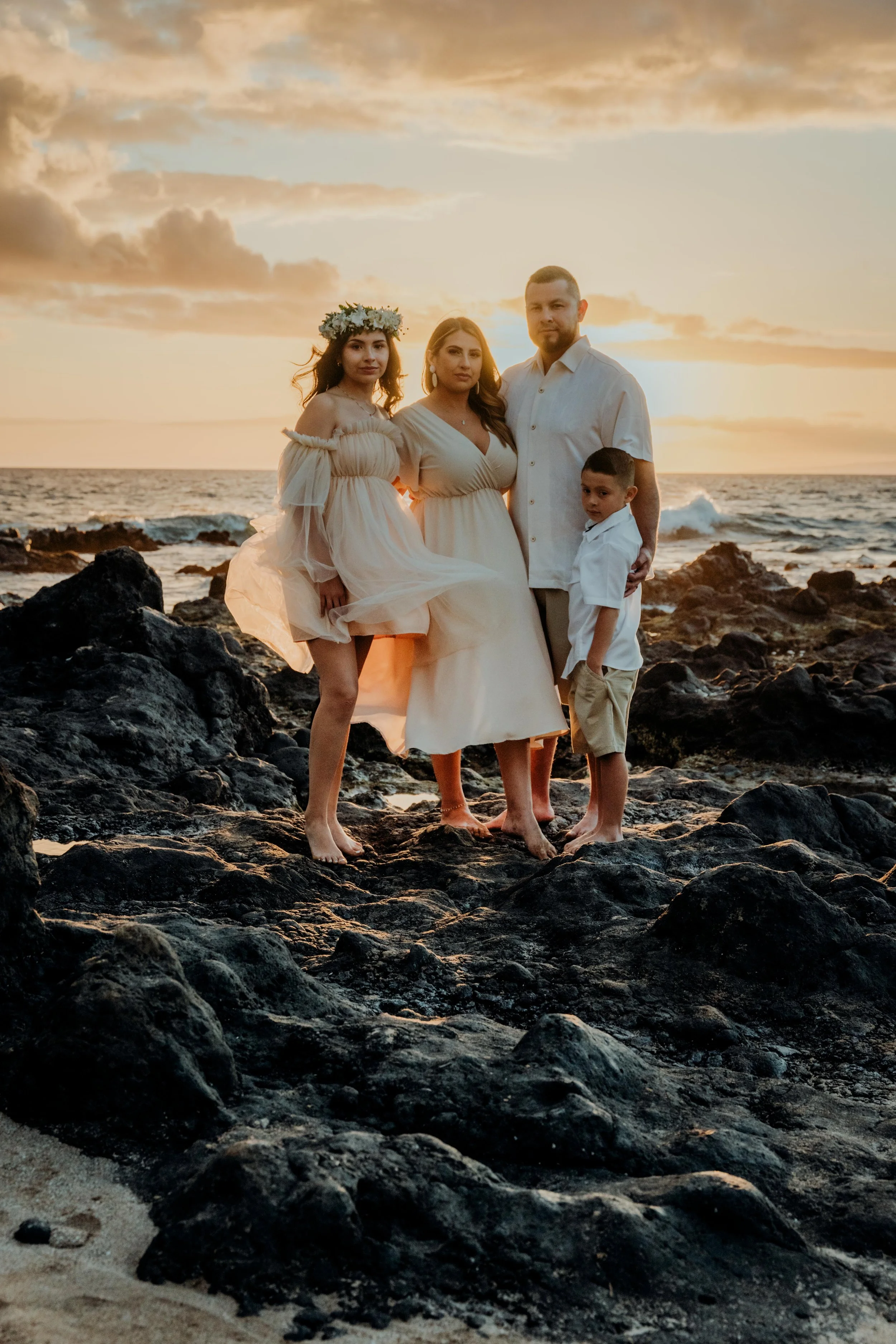 A family of four stands barefoot on rocky beach during sunset, dressed in white attire, with the ocean and cloudy sky in the background.