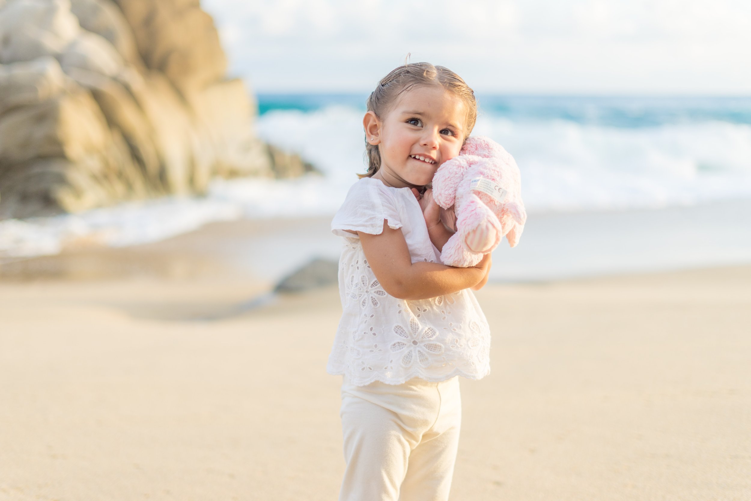 A young girl holding a pink stuffed animal on a beach with rocks and ocean waves in the background.