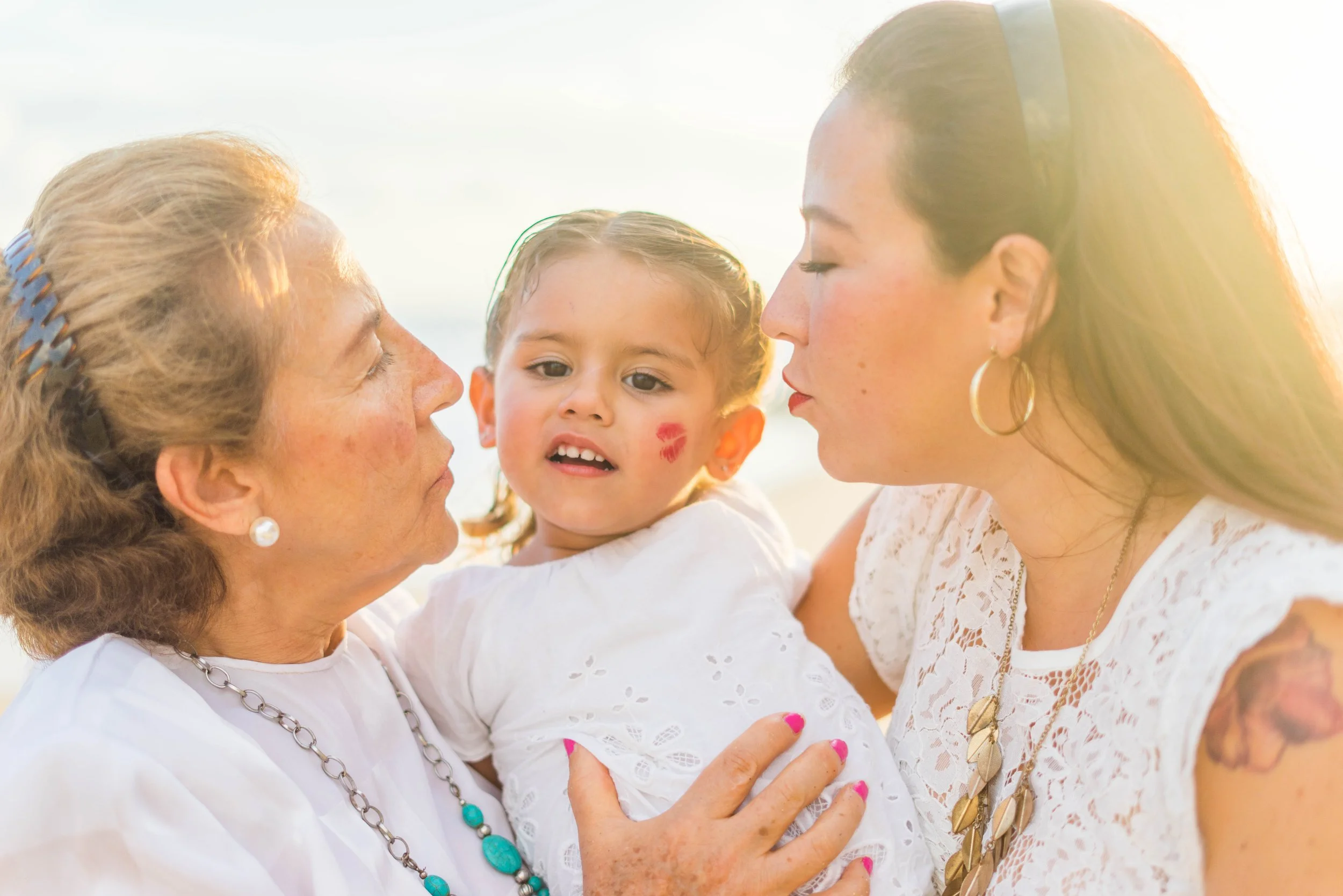 Two women and a young girl face-to-face outdoors, with the women about to kiss the girl on the cheeks. The girl has face paint on her cheek and is looking at the women. The women are wearing white tops and jewelry, and the girl is in a white dress.