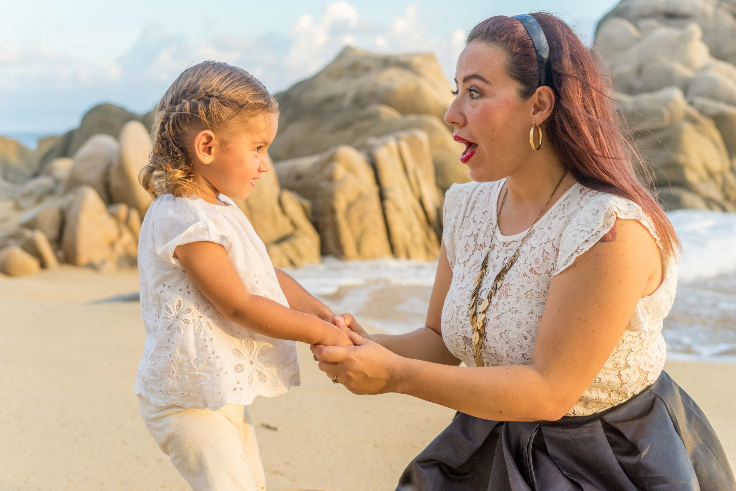 A woman and a young girl on a beach hold hands and face each other, with rocks and ocean waves in the background.