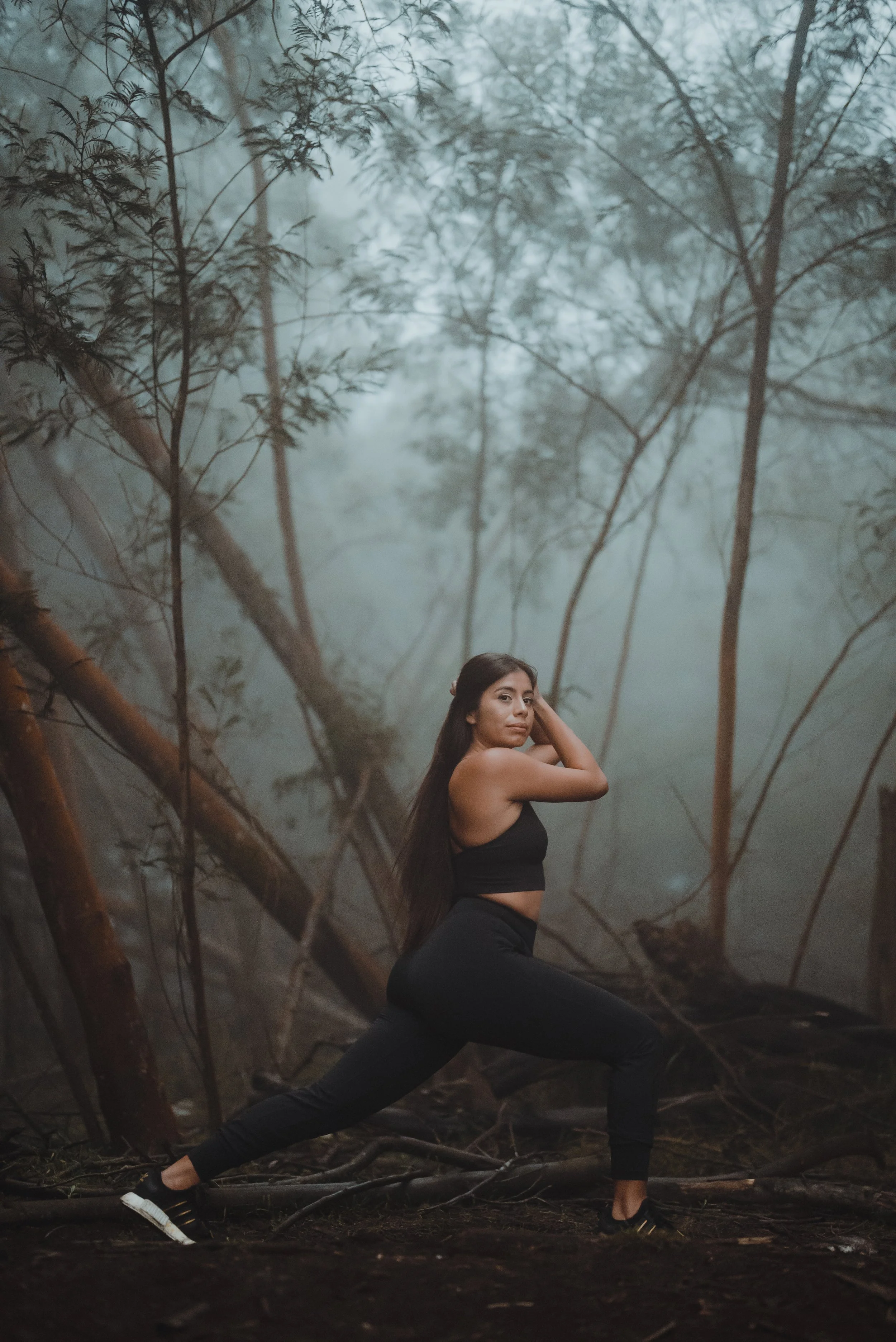 A woman in black athletic wear outdoors, performing a yoga stretch in a foggy forest with trees and roots on the ground.