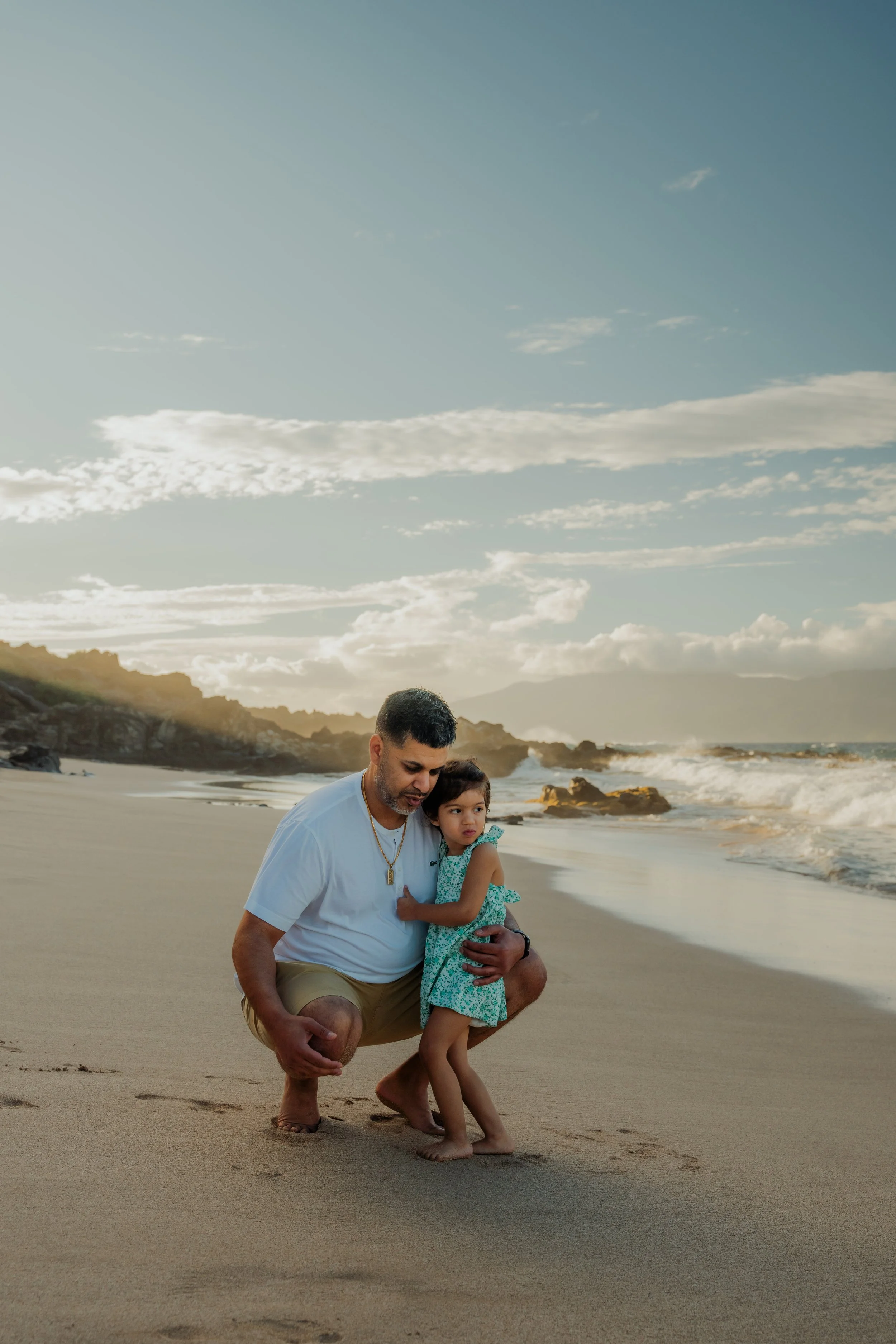 Man and young girl hugging on the beach with waves in the background and a partly cloudy sky.