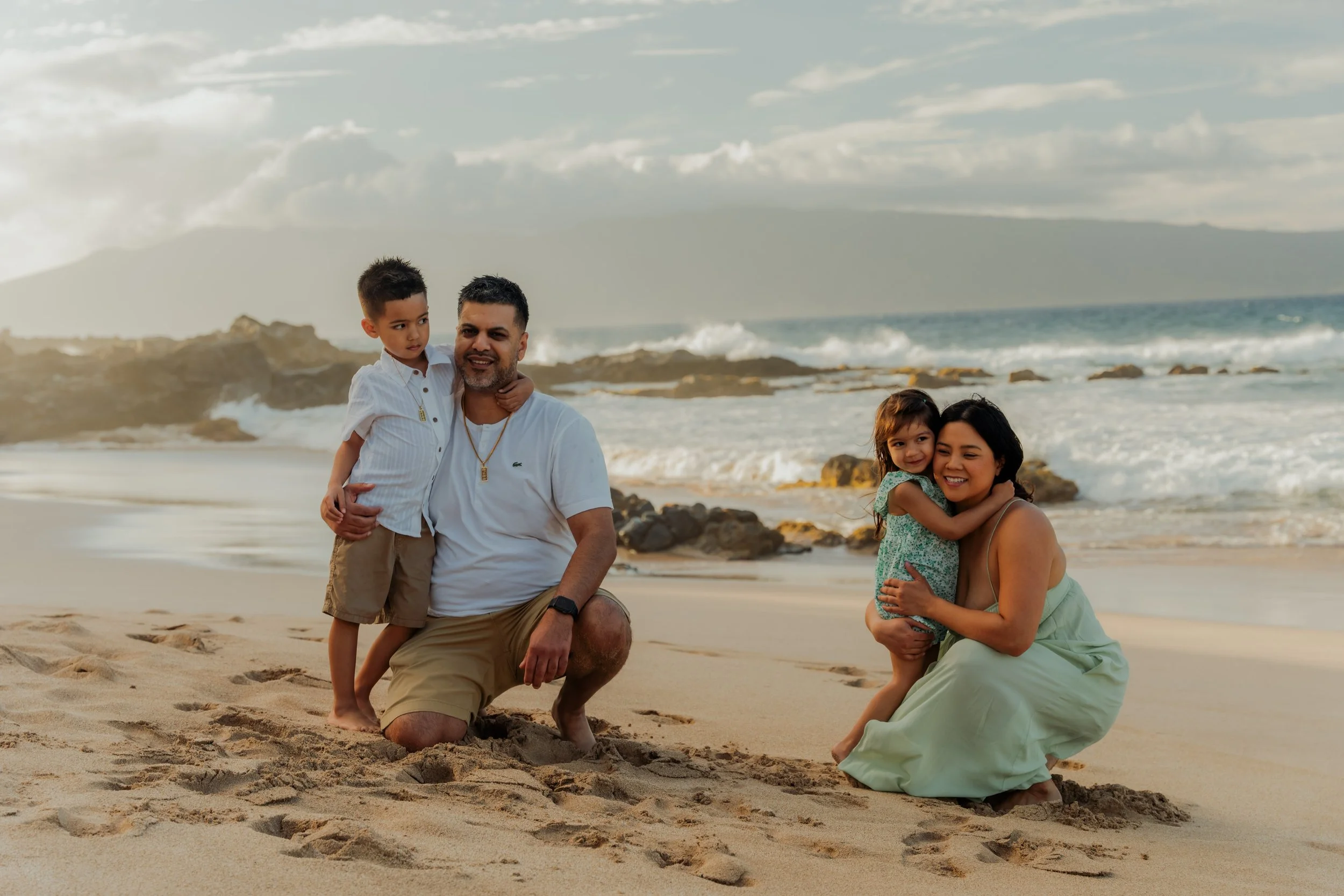A family of four enjoying time together on the beach, with the ocean and rocks in the background. The parents are kneeling in the sand, holding their children close, smiling and looking happy.
