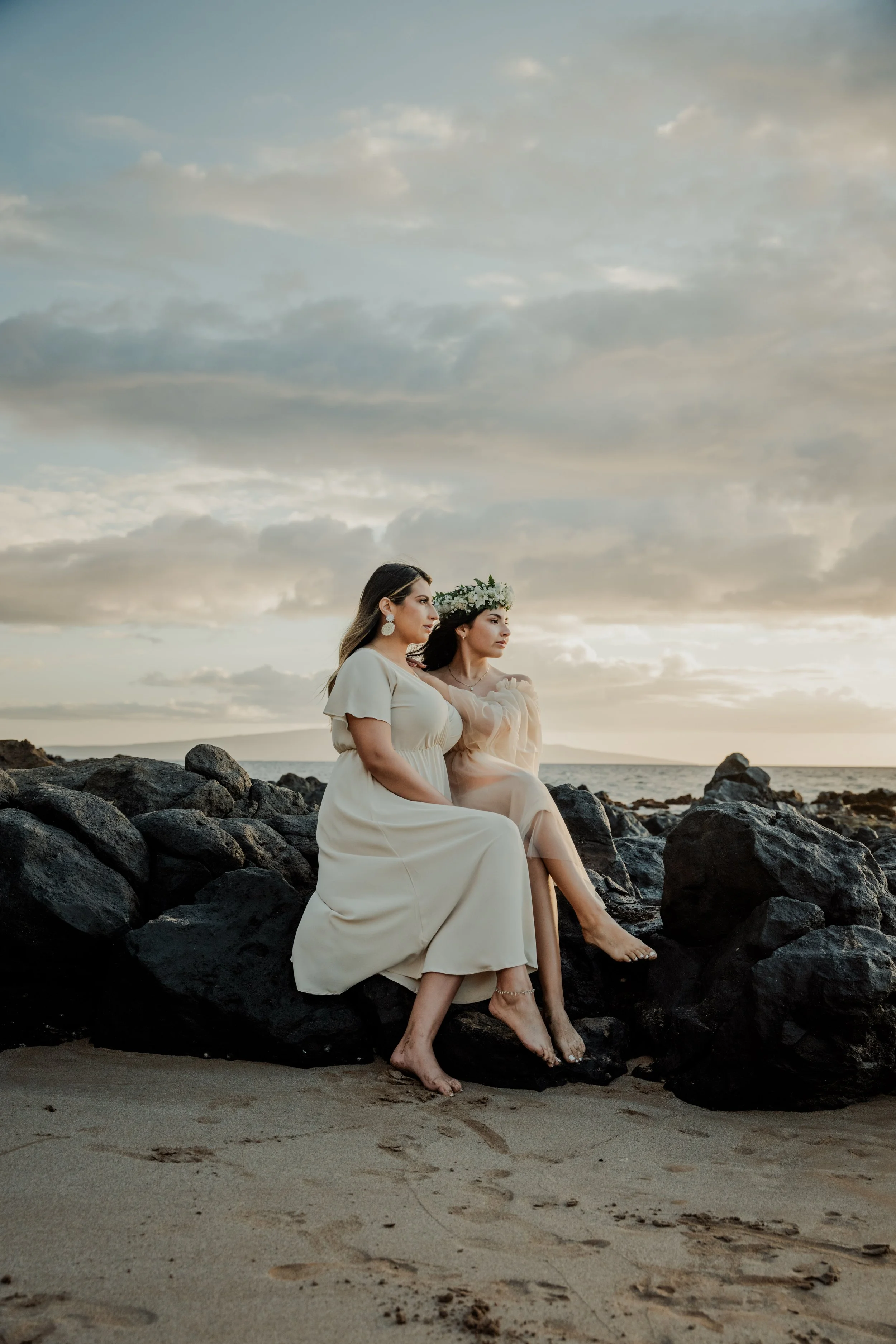 Two women dressed in light-colored dresses sitting on rocks at the beach during sunset, one wearing a flower crown and both barefoot.