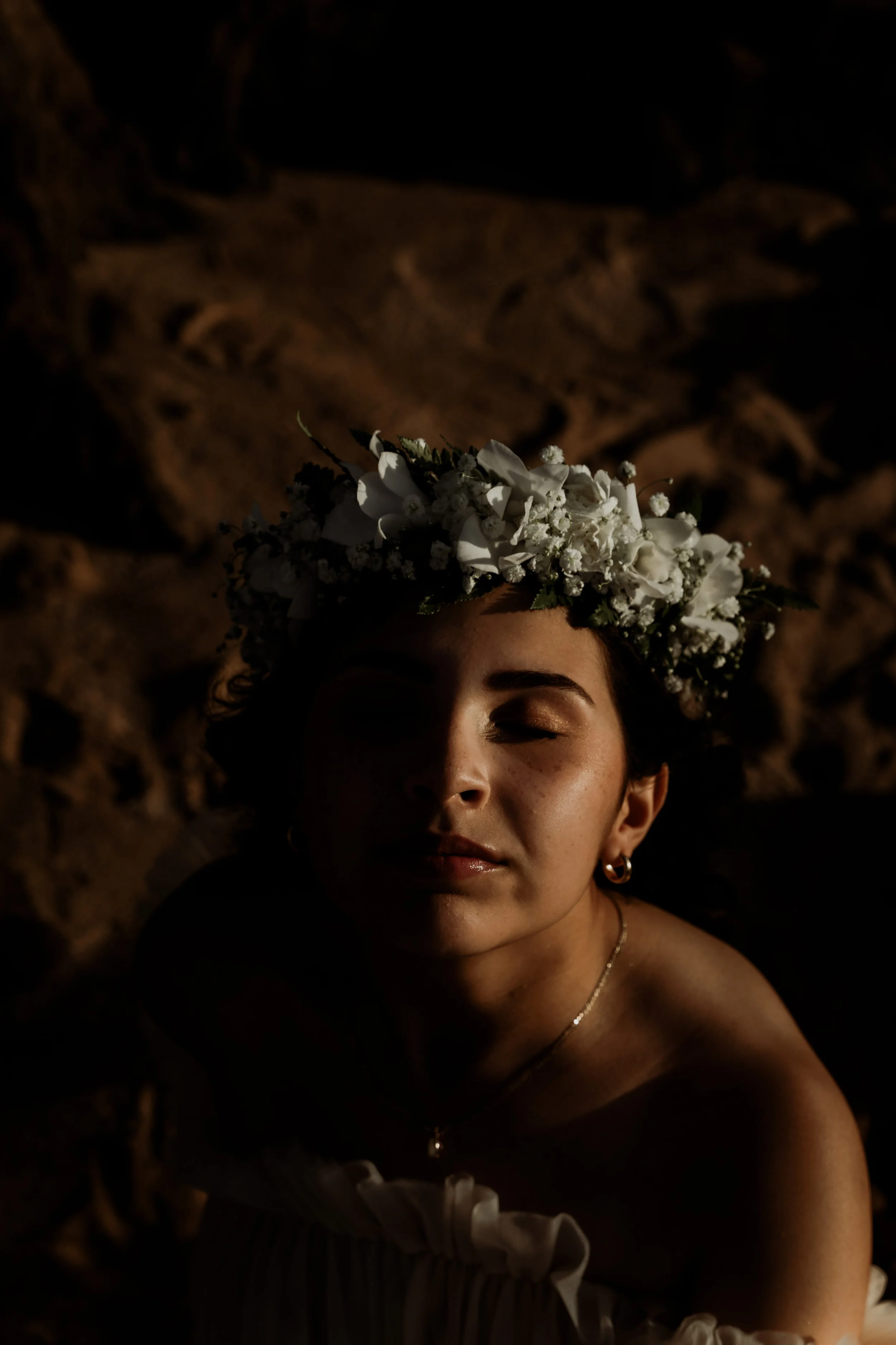 A woman with a floral crown on her head, eyes closed, in a dark setting with a rocky background.