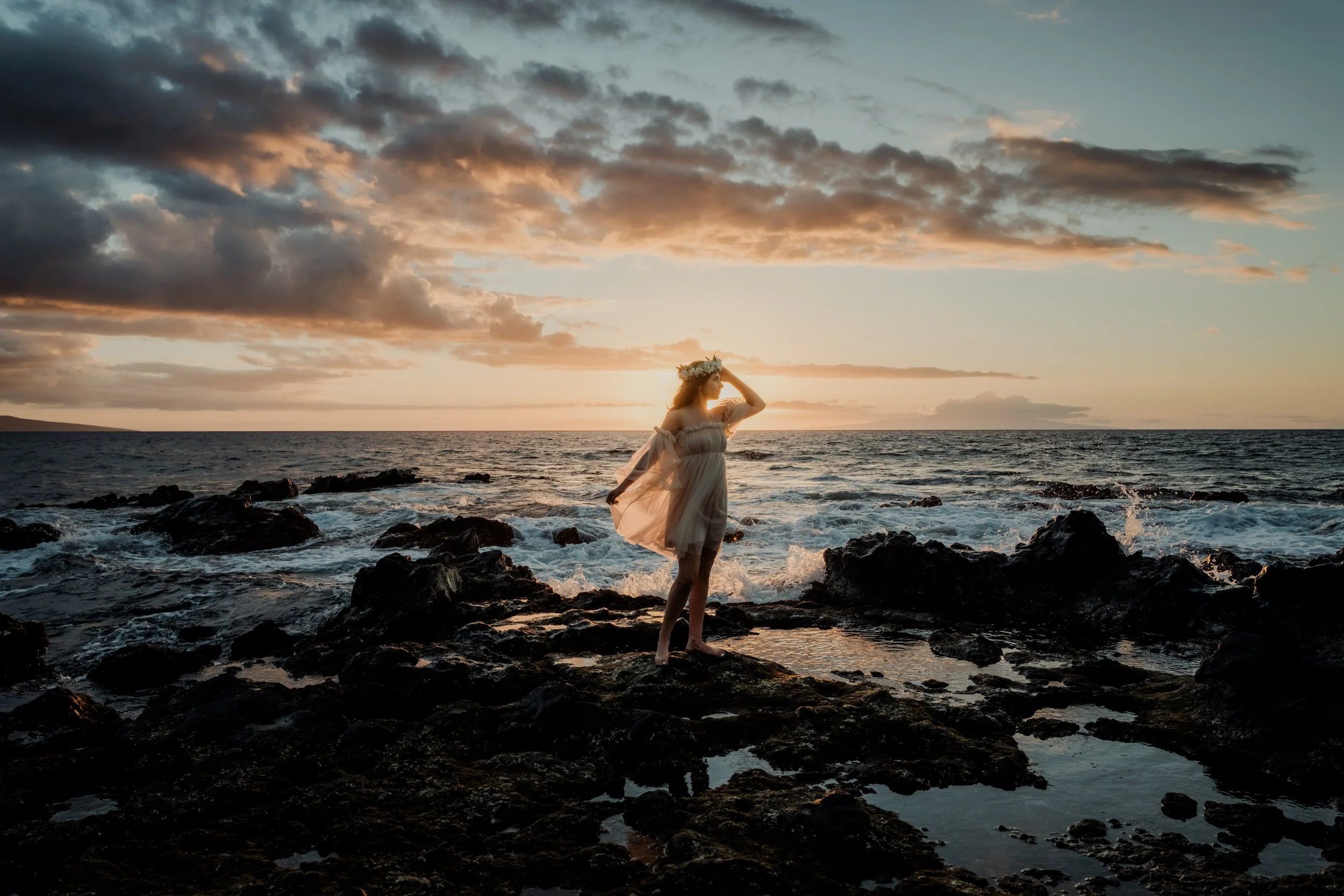 A woman in a flowing dress and floral crown standing on rocks by the ocean during sunset.