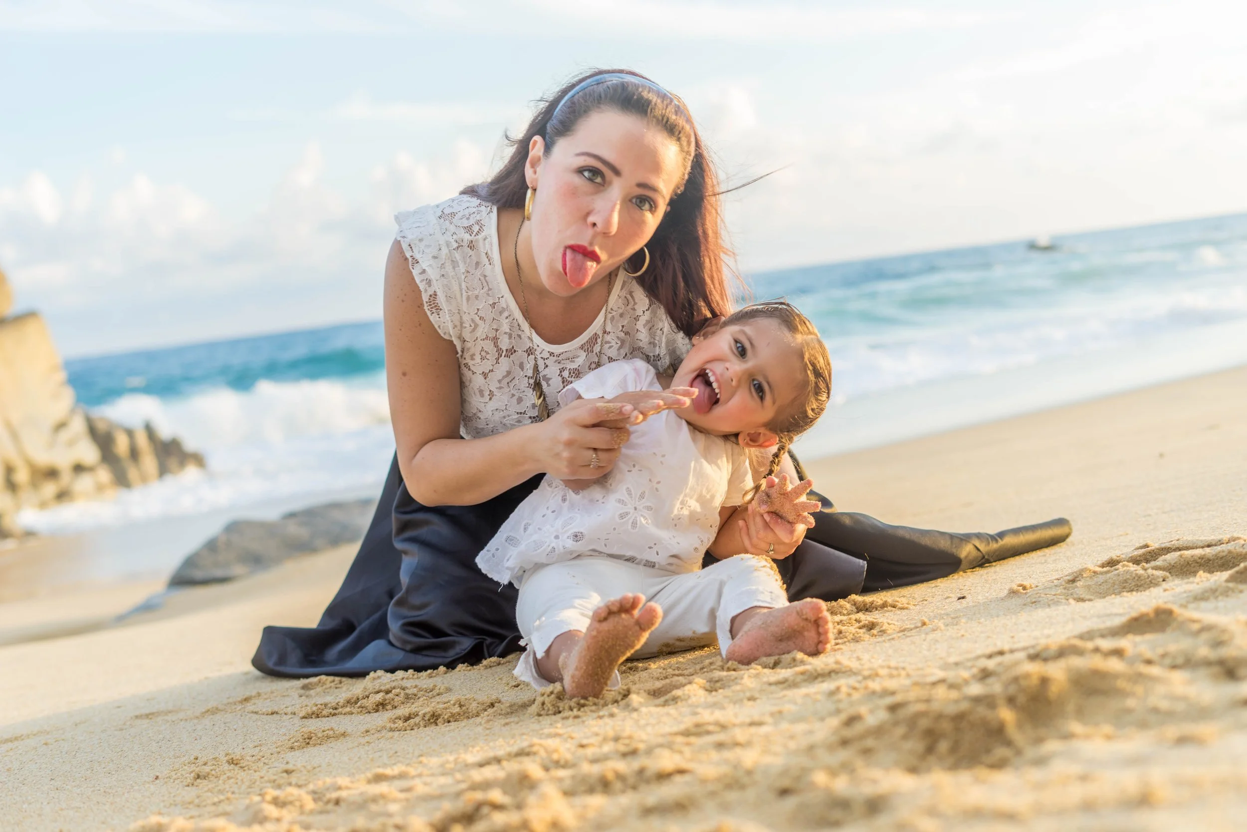 A woman and a young girl sitting on the sandy beach, playing with sand, with the ocean and blue sky in the background, both sticking their tongues out and making funny faces.