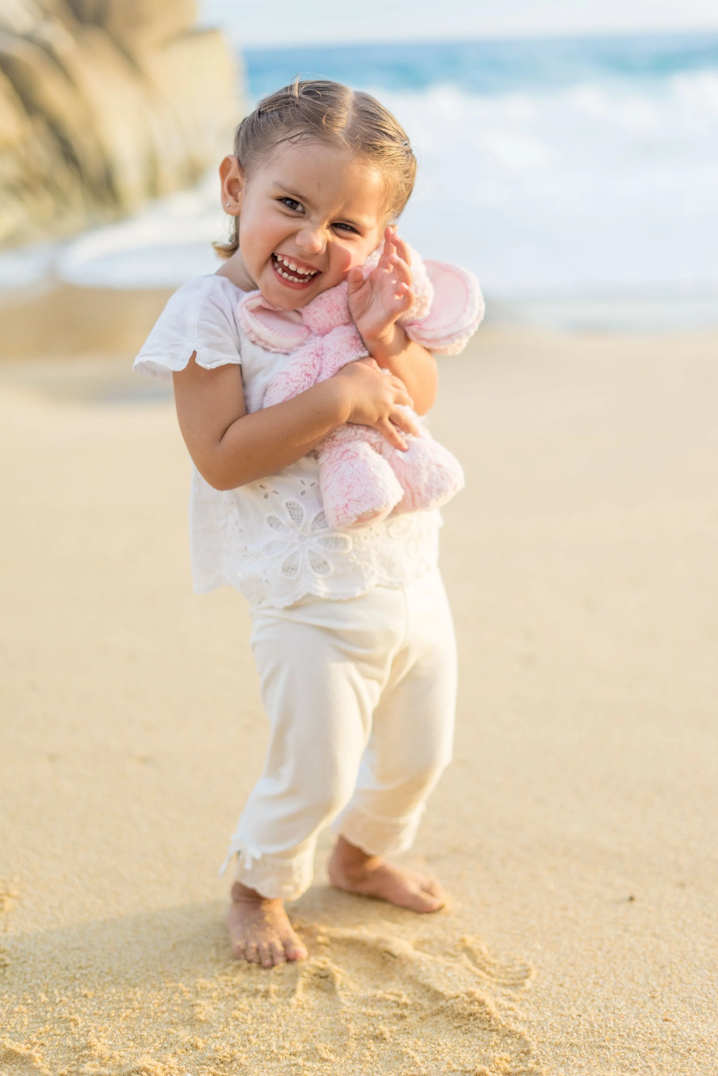 A smiling young girl hugging a pink stuffed animal on a sandy beach, with ocean waves in the background.