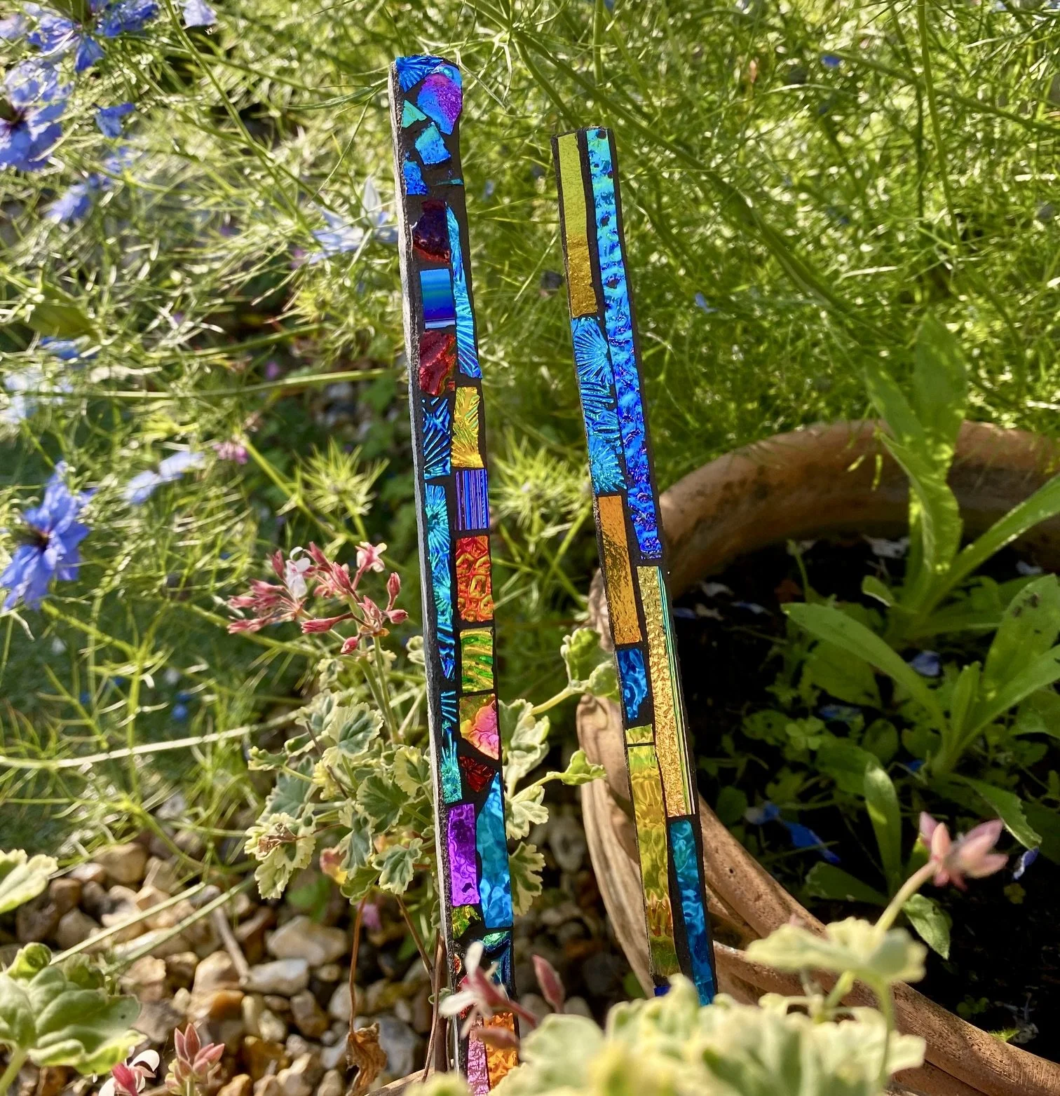 Two colorful stained glass-like glass bars standing upright among green plants and flowers in a garden.