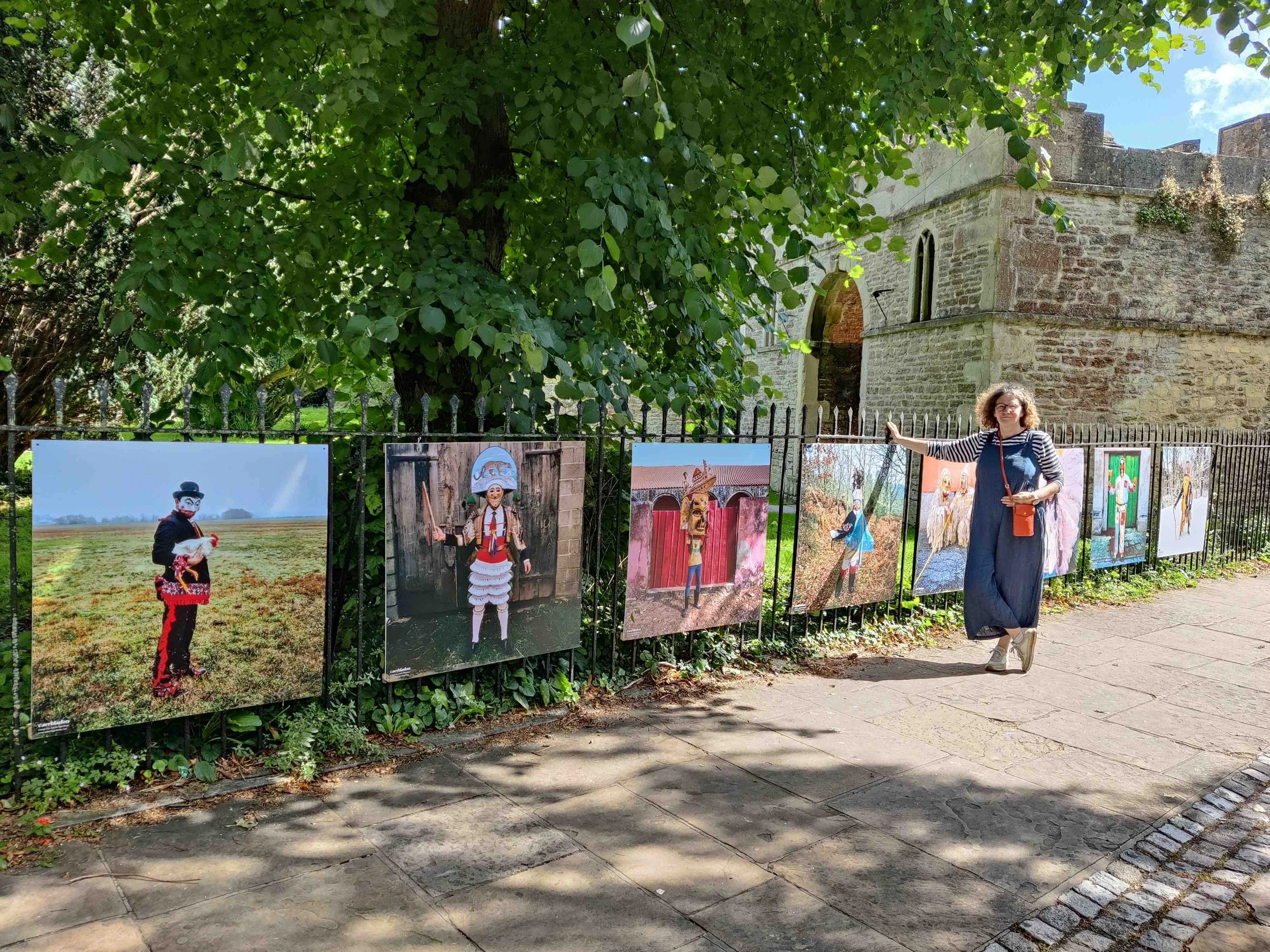 Karen Drake standing next to a colourful outdoor photo exhibit , with a historic stone building called Malmesbury Abbey and lush green tree in the background.