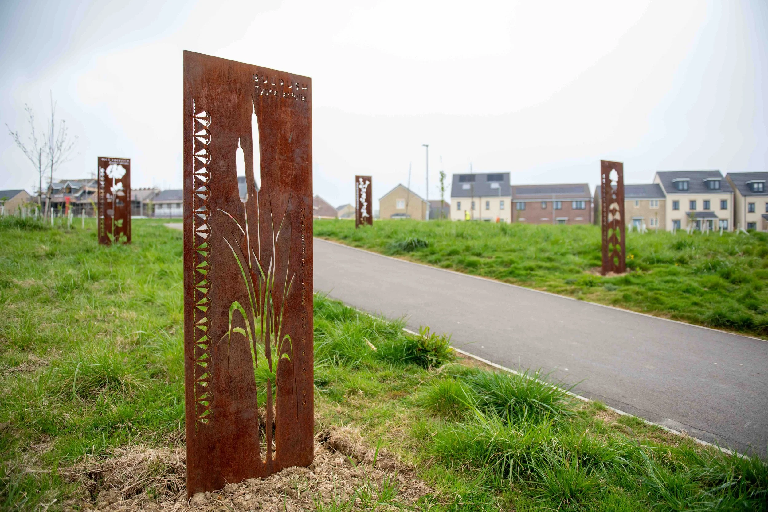 A pathway in a residential neighborhood with metal art panels depicting plants and natural scenes along the grass.