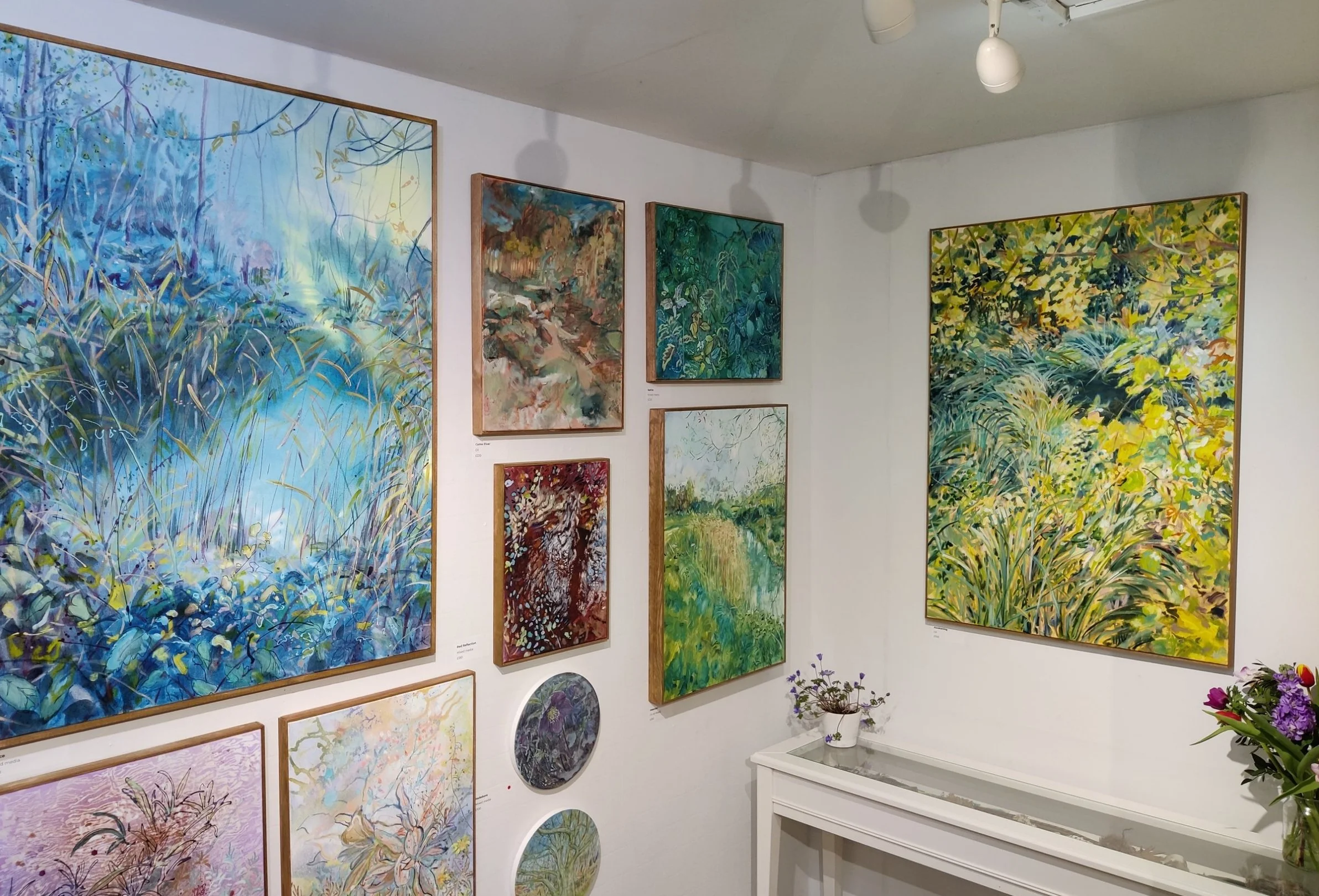 A gallery wall featuring multiple colorful landscape and nature paintings in various sizes. The paintings depict scenes of grass, trees, water, and foliage. There are also a few small circular artworks and a purple-flowered plant on a white table below the paintings.