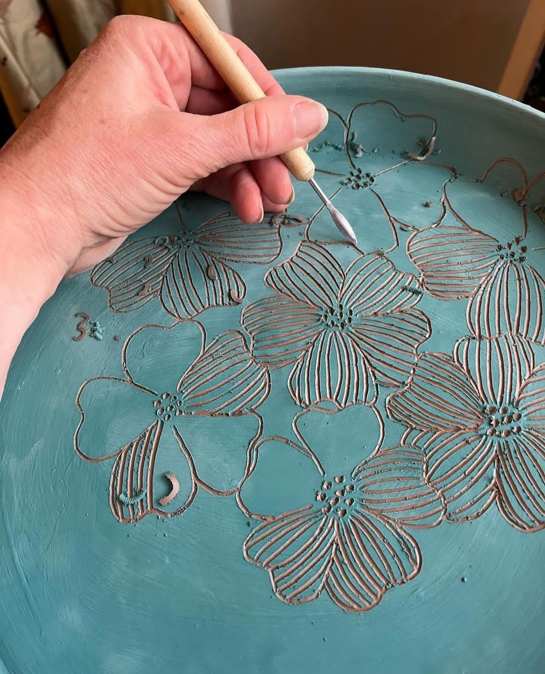 A person carving floral patterns into a light blue ceramic plate using a small carving tool.