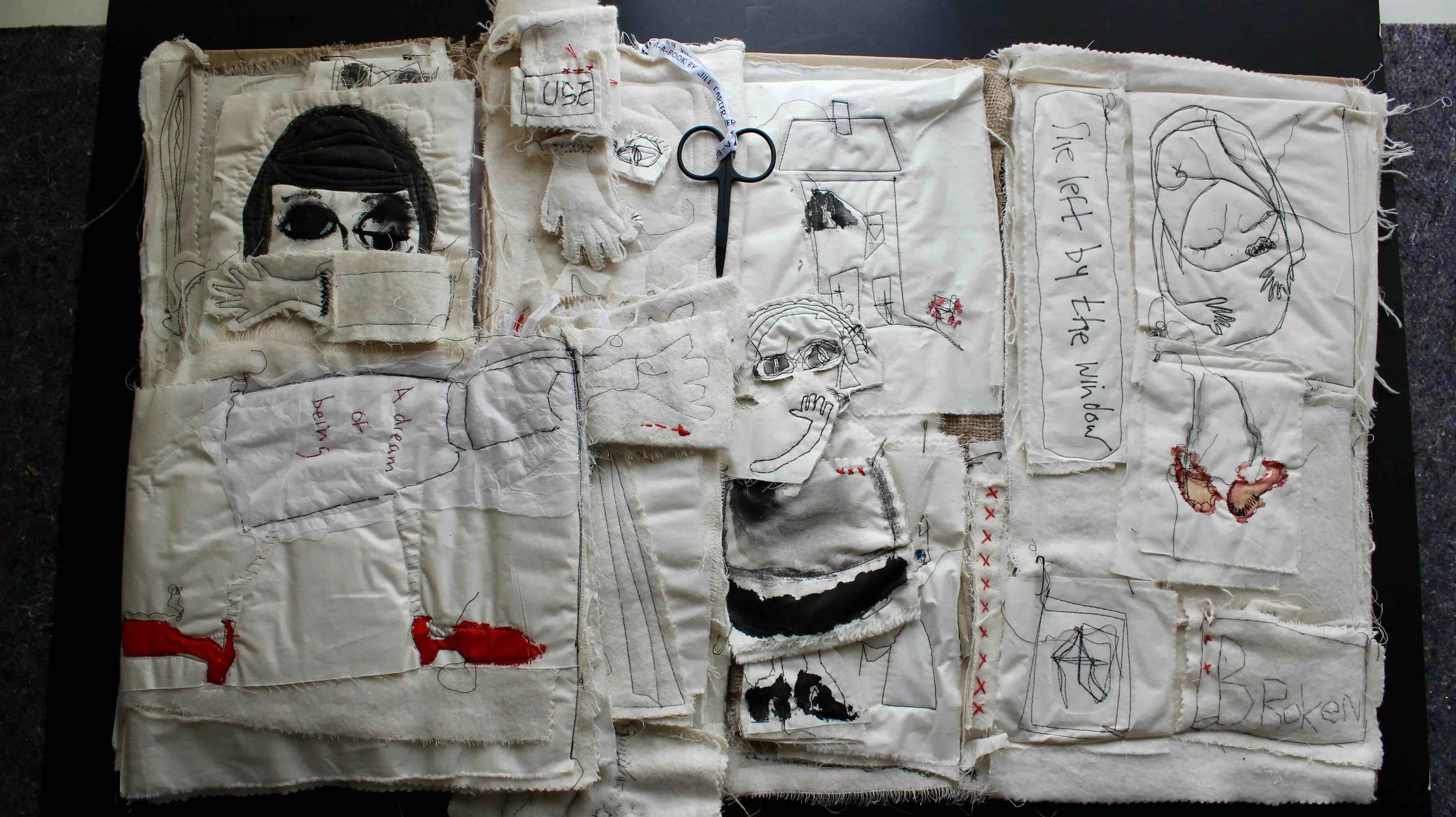 A textile art piece made of multiple fabric patches with embroidered, stitched, and drawn images and text, including a female face, a girl with glasses, a woman lying down, and various sketches, with some sections torn or frayed.