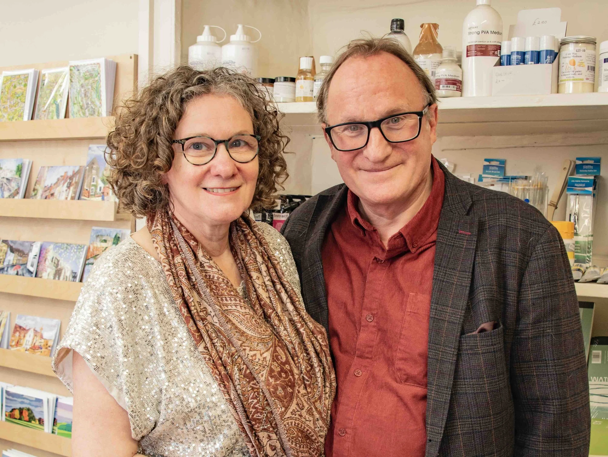 A smiling woman called Karen Drake with curly hair, glasses, and a patterned scarf stands next to a smiling man called David Drake with glasses, wearing a red shirt and a dark blazer, in Caerblad with books and art products on shelves behind them.