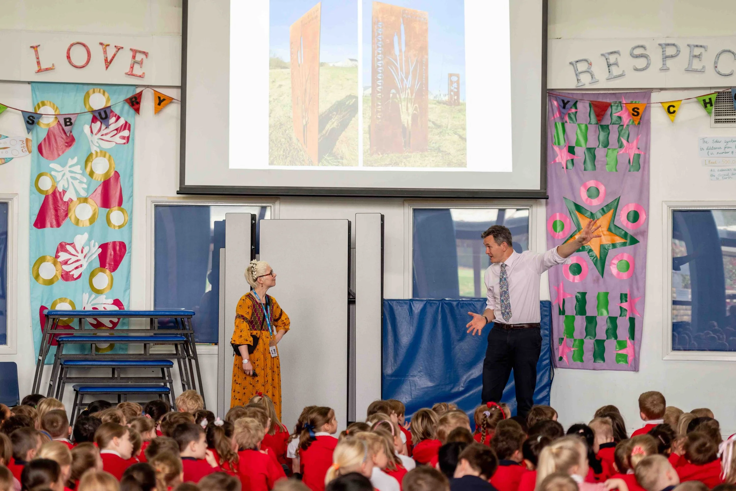 A classroom presentation with children in red uniforms.  The Head is gesturing toward a screen displaying outdoor art. Artist Kerry Lemon in a yellow dress and glasses watches . Decorated walls with  banners and cutouts read 'Love' and 'Respect.'