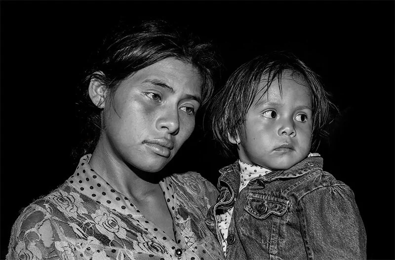 Black and white photo of a woman holding a young girl, both with serious expressions, against a dark background.