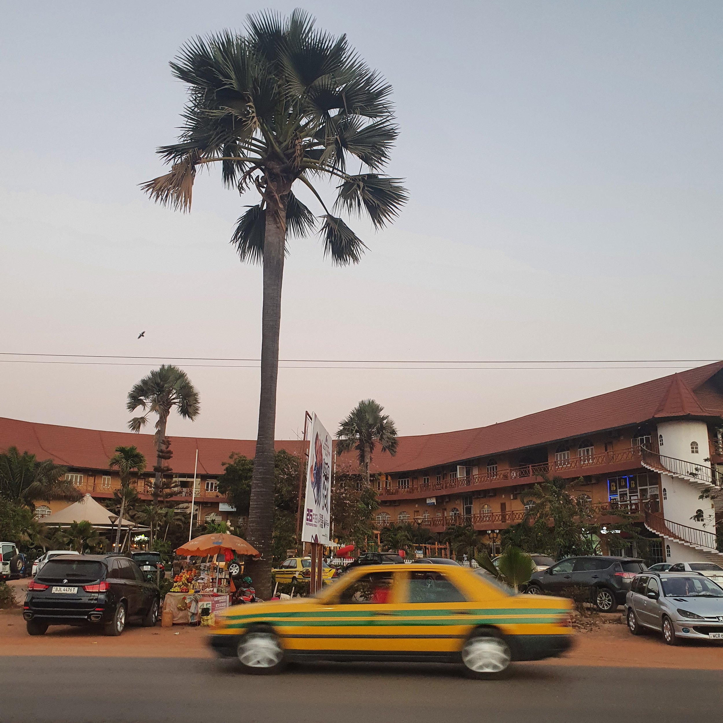 Street scene with a yellow and green taxi, parked cars, palm trees, a roadside vendor with an umbrella, a large building with a red roof, and a billboard, under a clear sky.