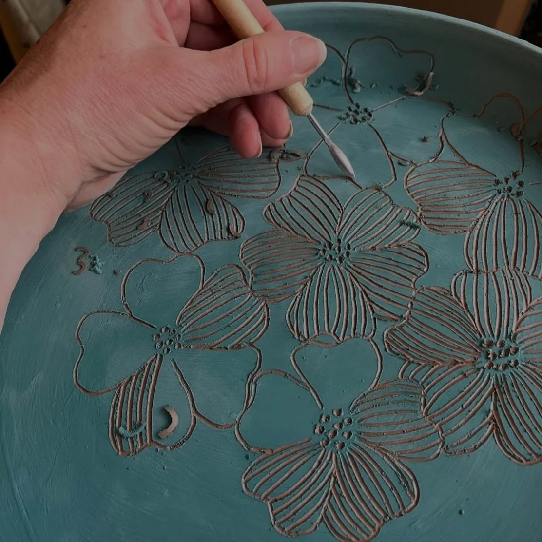 Close-up of a hand carving floral patterns into a blue ceramic piece using a carving tool.