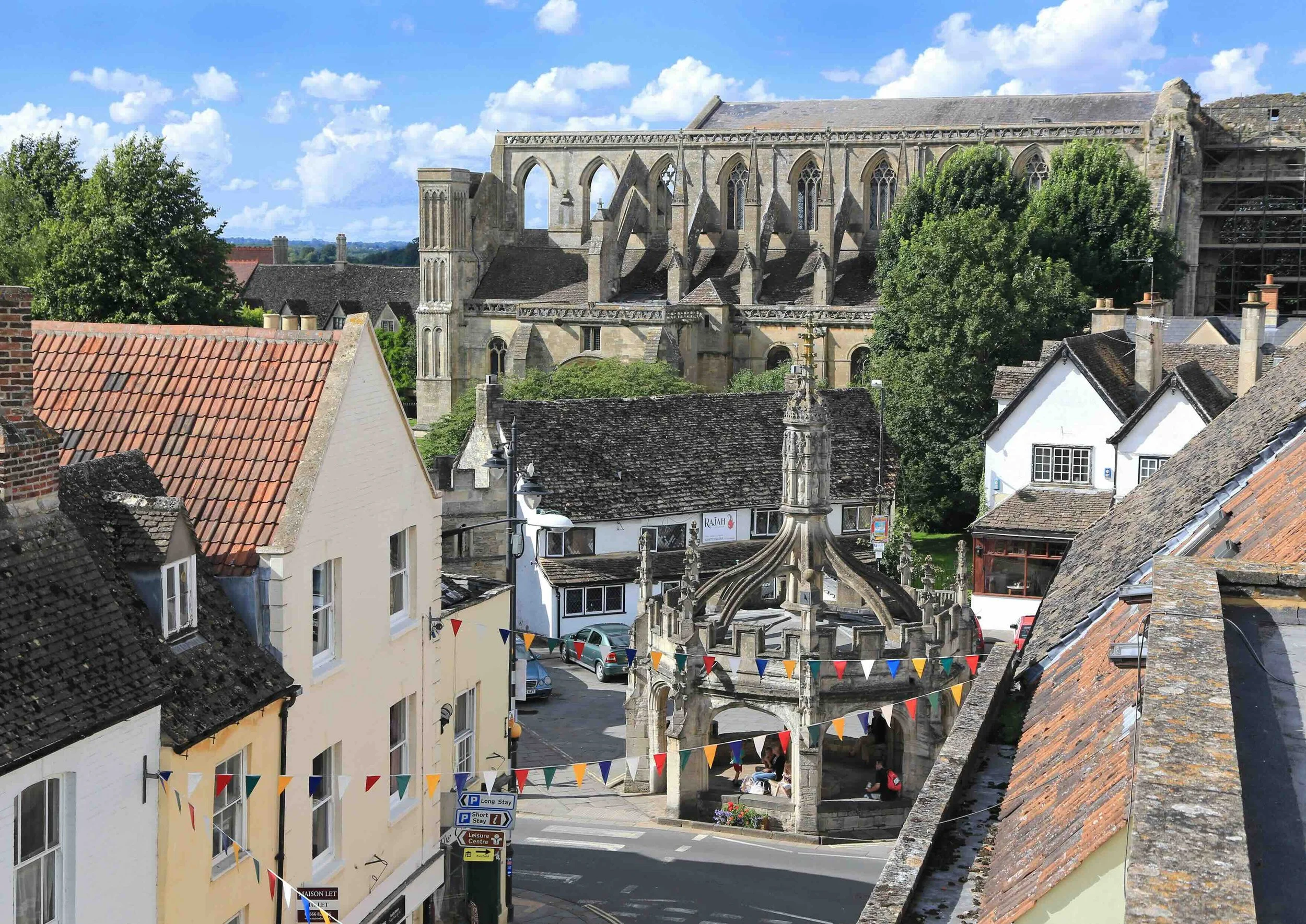 View of historic Malmesbury Abbey, surrounded by smaller buildings with tiled roofs. The stone Market Cross in the foreground. Lush green trees and a bright blue sky with fluffy white clouds are also visible.