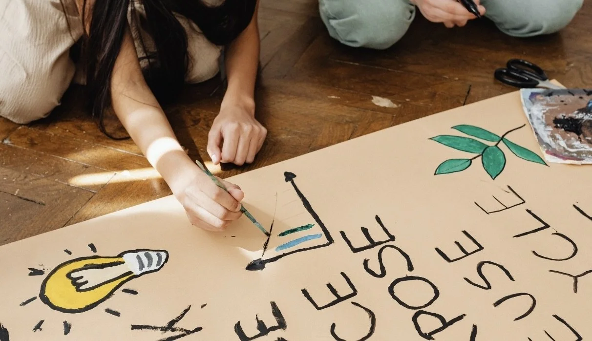 Person lying on the floor, painting a large poster with a lightbulb, a sun, and leafy branches, with the words "The best house of the youth" written on it.