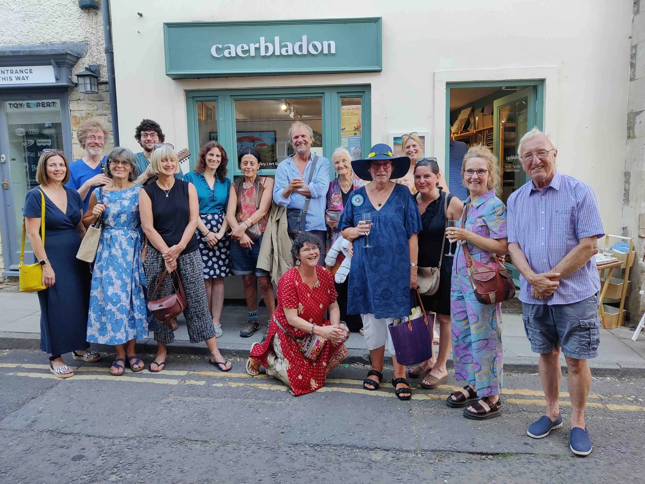 Group of 16 local artists gathered outside art gallery named Caerbladon, posing for a photo, some holding glasses, dressed casually with a woman in a large sun hat, in a lively outdoor setting.