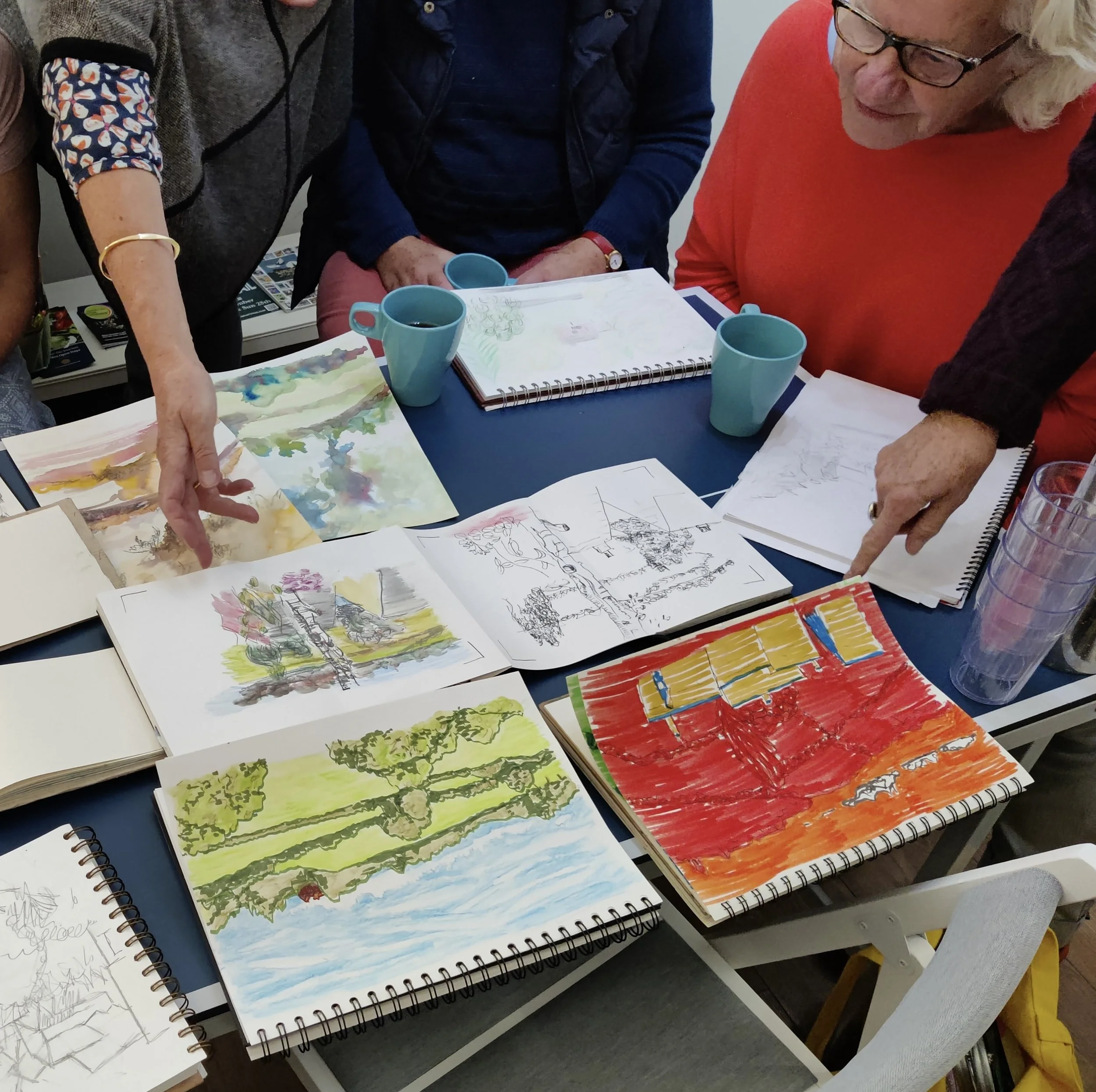 Several people gathered around a table covered with open sketchbooks and watercolor paintings, discussing and pointing at the artwork. There are blue cups on the table.
